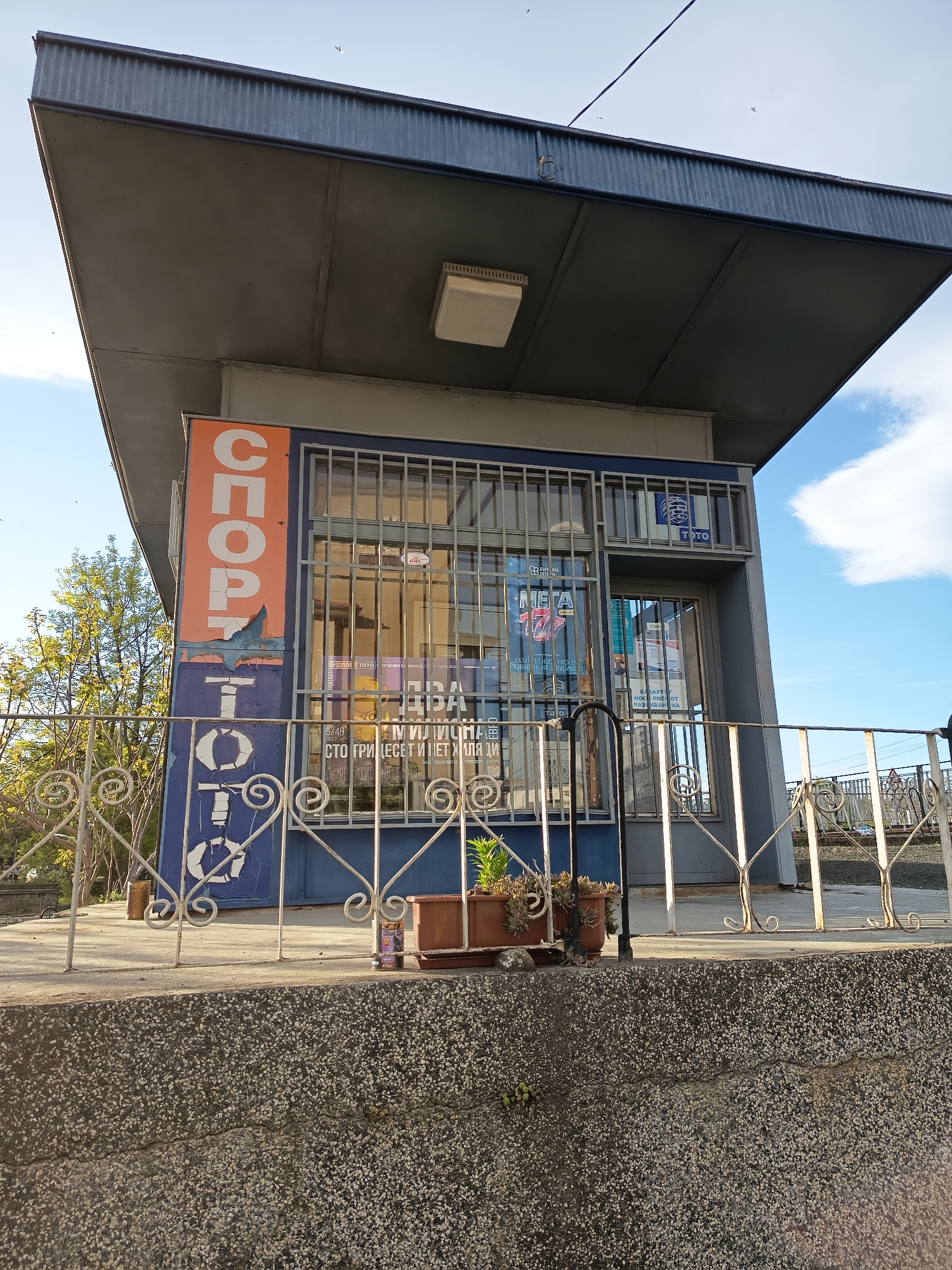A small, blue and orange kiosk, likely a lottery or betting shop, stands under a grey, overhanging roof. The structure is fortified with metal bars over its windows and door. To the left, an orange sign with peeling paint reads "СПОРТ" (SPORT) vertically, transitioning to "ТОТО" below, indicating "Sport Toto." Inside the kiosk, various posters are visible through the windows, including a "TOTO" logo, "МЕГА 777" (MEGA 777), and phrases in Cyrillic like "6/49," "МИЛЛИОНА" (MILLIONA - millions), and "СТО ГРИДЕСЕТ И ПЕТ ХИЛЯДИ" (one hundred thirty-five thousand, likely a prize amount). Other partially visible text includes "КАЗАН" (Kazan - possibly winnings) and "РАЗВИВАН НА" (developing/expanding).
In front of the kiosk is a white, ornate metal fence, behind which sit several brown potted plants. The foreground features a textured, grey concrete wall or ledge. The scene is illuminated by daylight, suggesting a clear, sunny day with some white clouds visible in the blue sky. The general absence of activity suggests the kiosk might be closed or captured during a quiet moment. Trees with green foliage are visible in the background to the left. The Cyrillic text on the kiosk indicates an Eastern European setting.