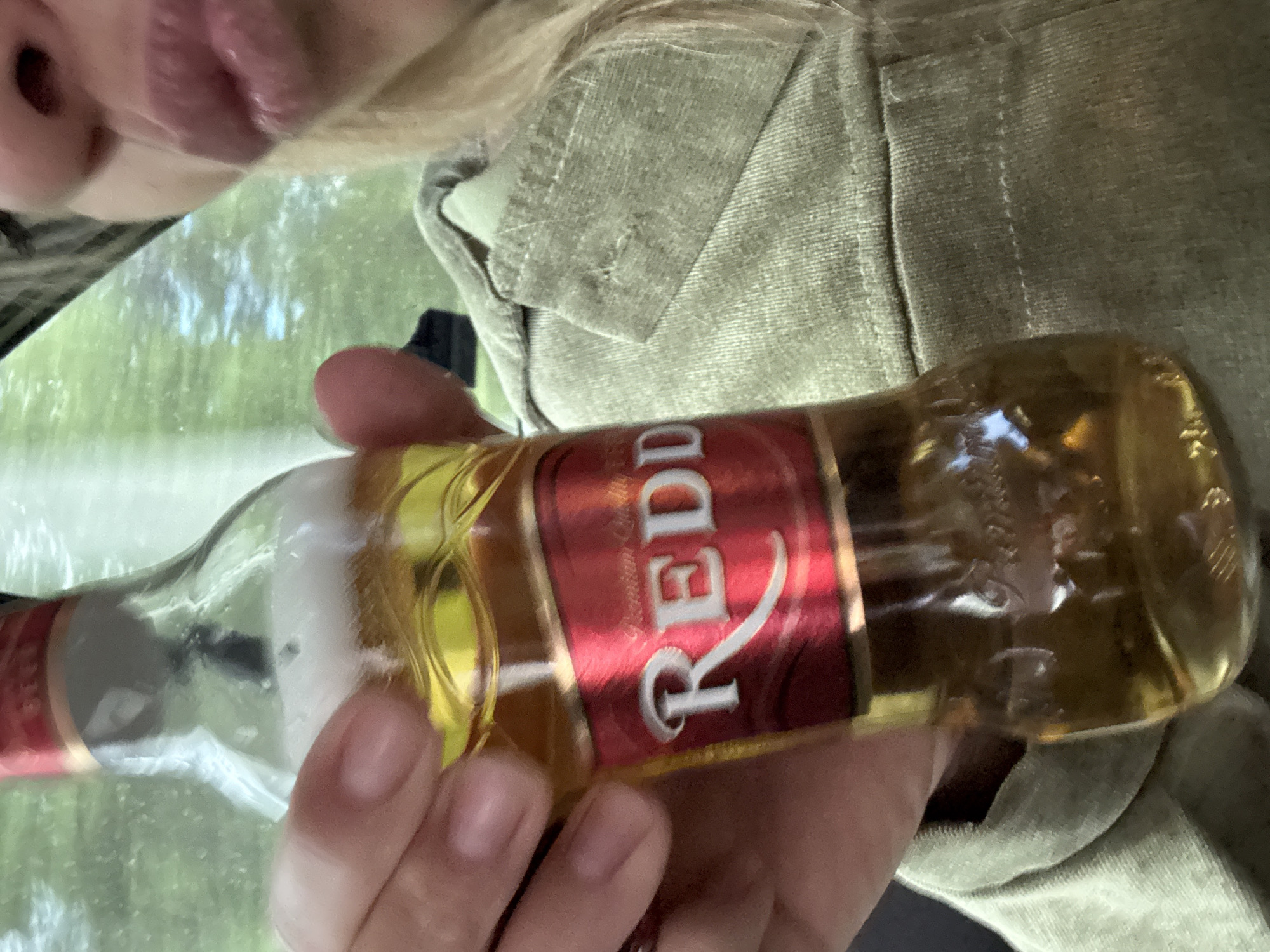 A person is holding a small bottle of Redd's beer. The person is wearing a light green jacket and has blonde hair. The bottle is mostly full and has a red label with "REDD" written in white letters. The background appears to be a blurry outdoor scene with greenery. There is no identifiable information to suggest this image is located in Zheleznodorozhny, Russia.
