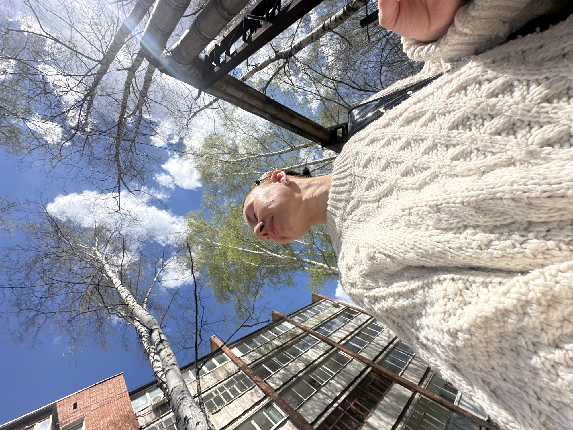A low-angle shot presents a person wearing a chunky, textured cream-colored sweater, positioned against a backdrop of a clear blue sky dotted with fluffy white clouds. Above the person, there's a network of bare tree branches and metal pipes. A building with multiple windows and a brick facade is partially visible at the bottom. The location is likely Votkinsk, Russia, based on the image's origin.