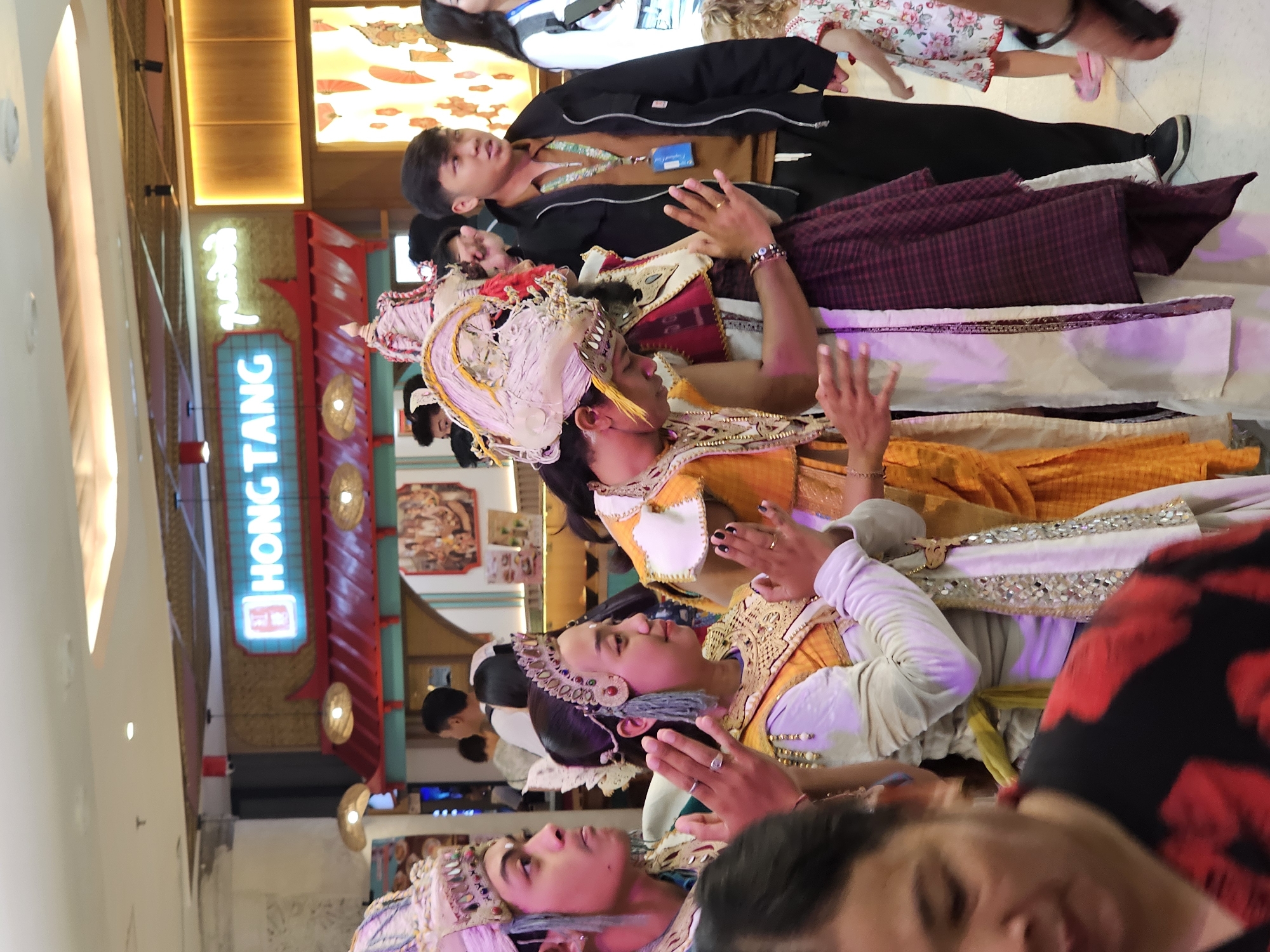 The image depicts a group of people in traditional Balinese attire, likely a performance or cultural event, inside a modern indoor setting resembling a shopping mall or cultural center. The individuals in the foreground are adorned with elaborate headdresses and ornate costumes, some with sequins and decorative trim, indicative of Balinese dance or ceremonial dress. Two individuals in the center are holding their hands together in a prayer-like gesture. Behind them, other participants and onlookers are visible. In the background, a brightly lit sign for "HONG TANG" is prominently displayed, suggesting a restaurant or business. The overall environment appears to be a well-lit, public space with elements of both traditional and contemporary design. The presence of Balinese cultural attire within a mall setting indicates a possible event or exhibit in Denpasar, Indonesia, aiming to showcase local culture to visitors or residents.
