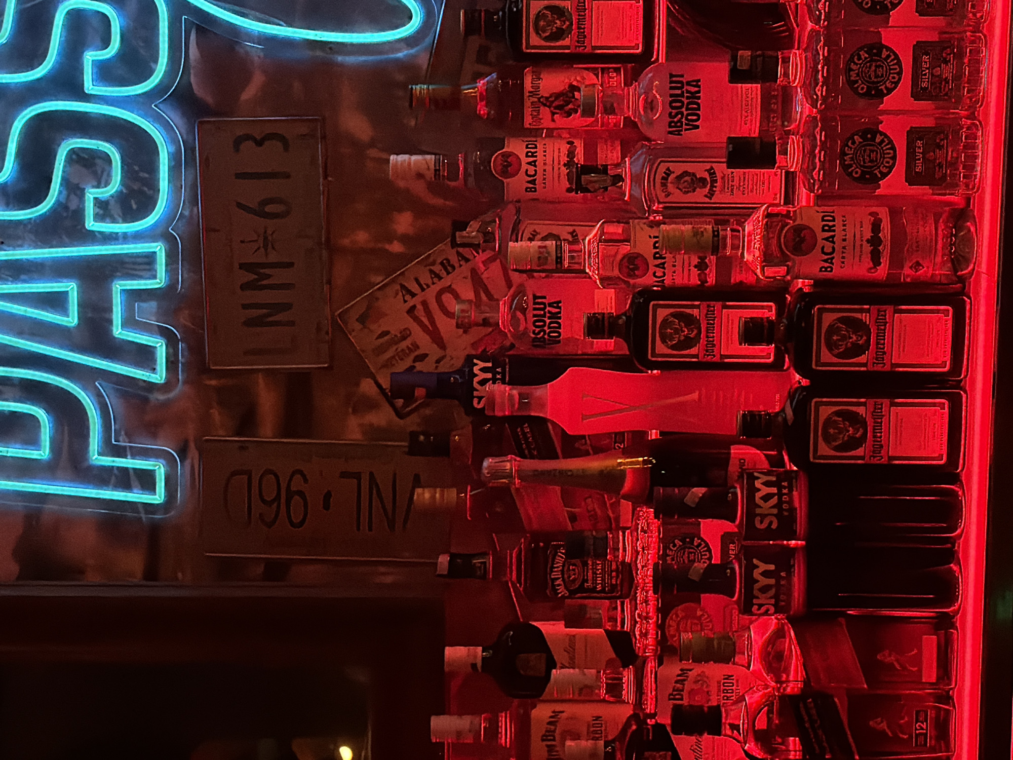 This image depicts a bar setting. The foreground features rows of liquor bottles including brands like Jim Beam, Jack Daniel's, Skyy Vodka, Jagermeister, Absolut Vodka, Bacardi, Captain Morgan, and Olmeca Tequila. Positioned behind the bottles are several license plates; one plate reads "NM-613," and another reads "/NL 96D". A vintage Alabama Veteran sign is also visible. To the left of the image, a bright neon sign spelling "PASS" is illuminated in a vibrant turquoise color. The bottles are lit with a strong red light, which creates a distinct ambiance. Given the information provided is limited, the location cannot be confirmed to be Chongqing High-tech Zone, China.