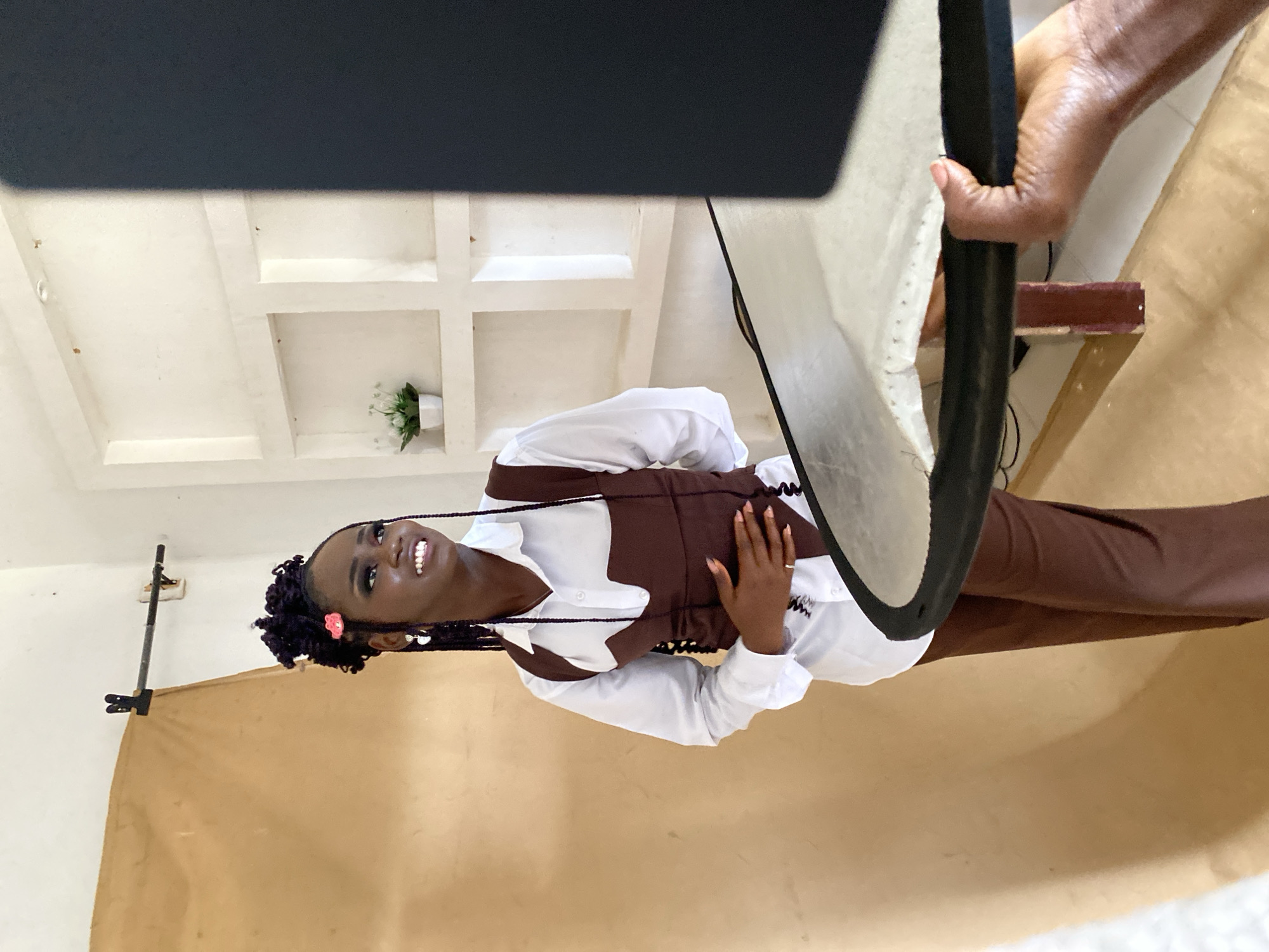 This media file features a young Black woman posing for a photo shoot in a studio setting. She has dark skin, braided dark purple hair adorned with a small pink flower, and is smiling broadly. She is wearing a white collared shirt with a dark brown corset or vest over it, and brown pants. Her left hand is resting on her waist, and a ring is visible on her finger.
The background of the studio consists of a plain beige backdrop. In the upper left of the image, a portion of a large black photography light or modifier is visible, and a hand is adjusting a white reflector or diffuser on the right side. The lighting suggests an indoor setup designed for photography. The overall mood is bright and positive, with the subject appearing joyful and confident. No specific time of day or weather conditions can be inferred, but the indoor setting implies controlled environmental conditions. The location context is Bukuru, Nigeria. There is no visible text in the image.