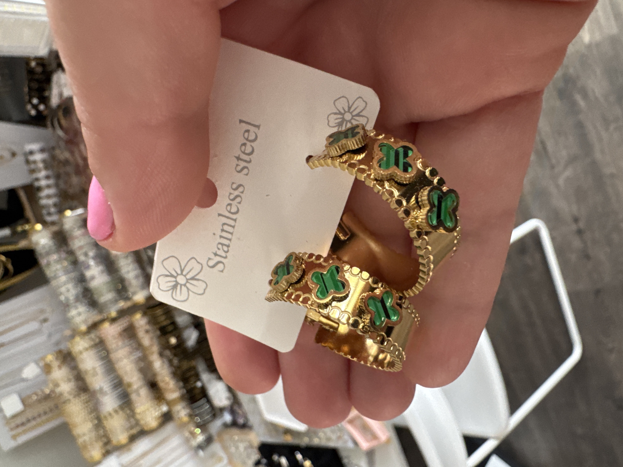 A person's hand, with a bright pink fingernail, holds a pair of gold hoop earrings adorned with green clover-like charms. The earrings are attached to a white card that reads "Stainless steel" in gray text, and features a simple line drawing of a flower. The background is blurred, showing a display of jewelry, likely in a retail setting in Wheeling, United States of America. The overall impression is a close-up shot of new earrings being presented or examined.
