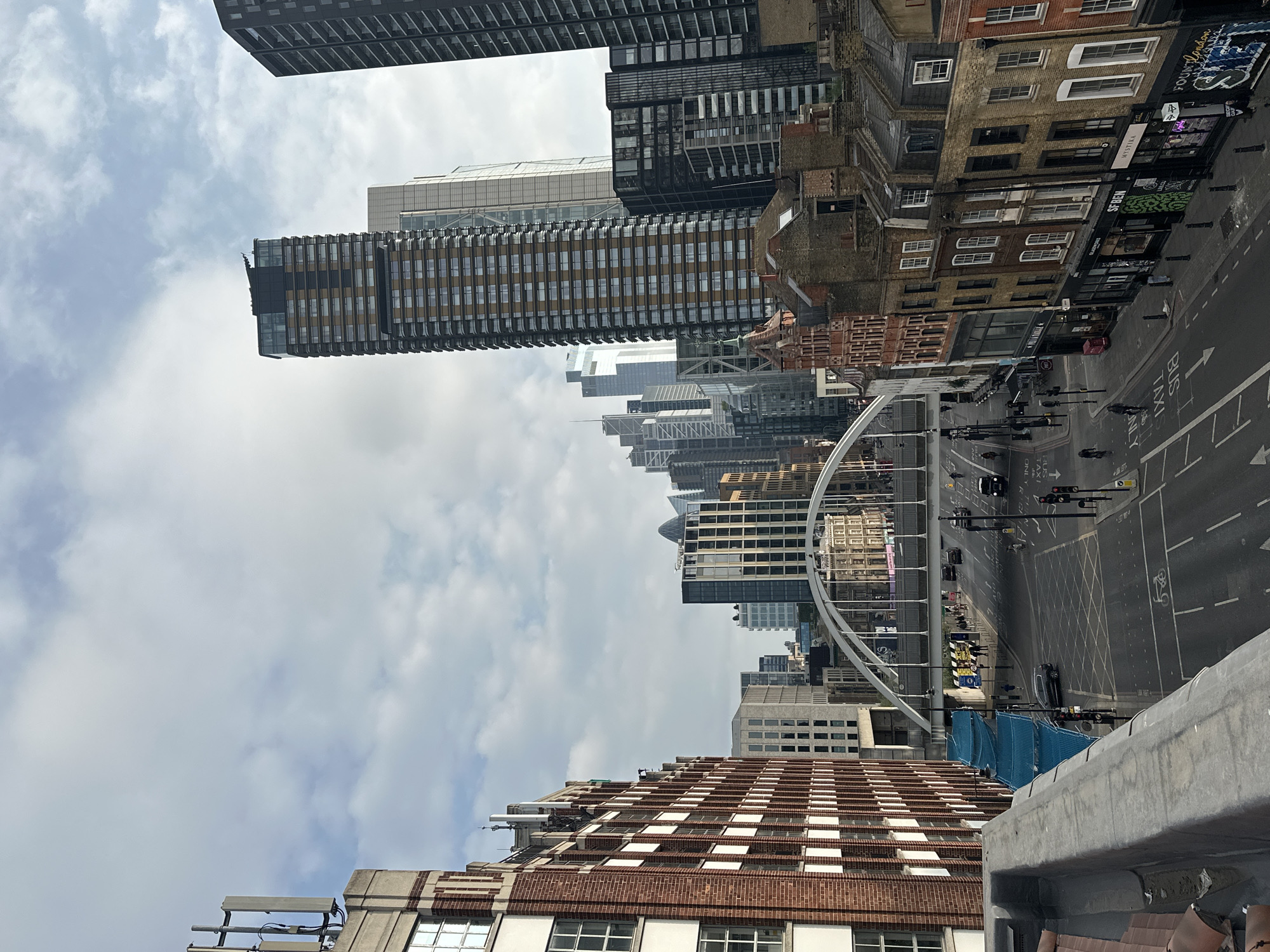 The image captures an urban landscape in London, United Kingdom. Tall buildings dominate the background, interspersed with modern skyscrapers and older brick structures. The sky is overcast with clouds. In the foreground, a bridge spans over a city street. Pedestrians walk along the sidewalks, and vehicles are visible on the road. The buildings along the street level house businesses, including shops with graffiti-covered storefronts.