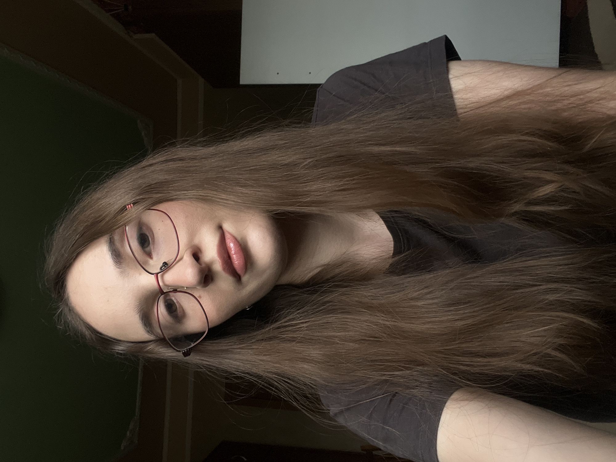 A medium shot shows a young woman with long, brown hair, wearing glasses and a dark t-shirt. She is looking at the camera, with her head tilted slightly to the left. Her lips are slightly parted and she is wearing a nude lipstick. The background is dark and out of focus, but appears to be an indoor setting. There is a white surface visible to the right of the woman, which might be a wall or a piece of furniture. The lighting is dim, with the light source seemingly coming from the front and slightly to the left.
