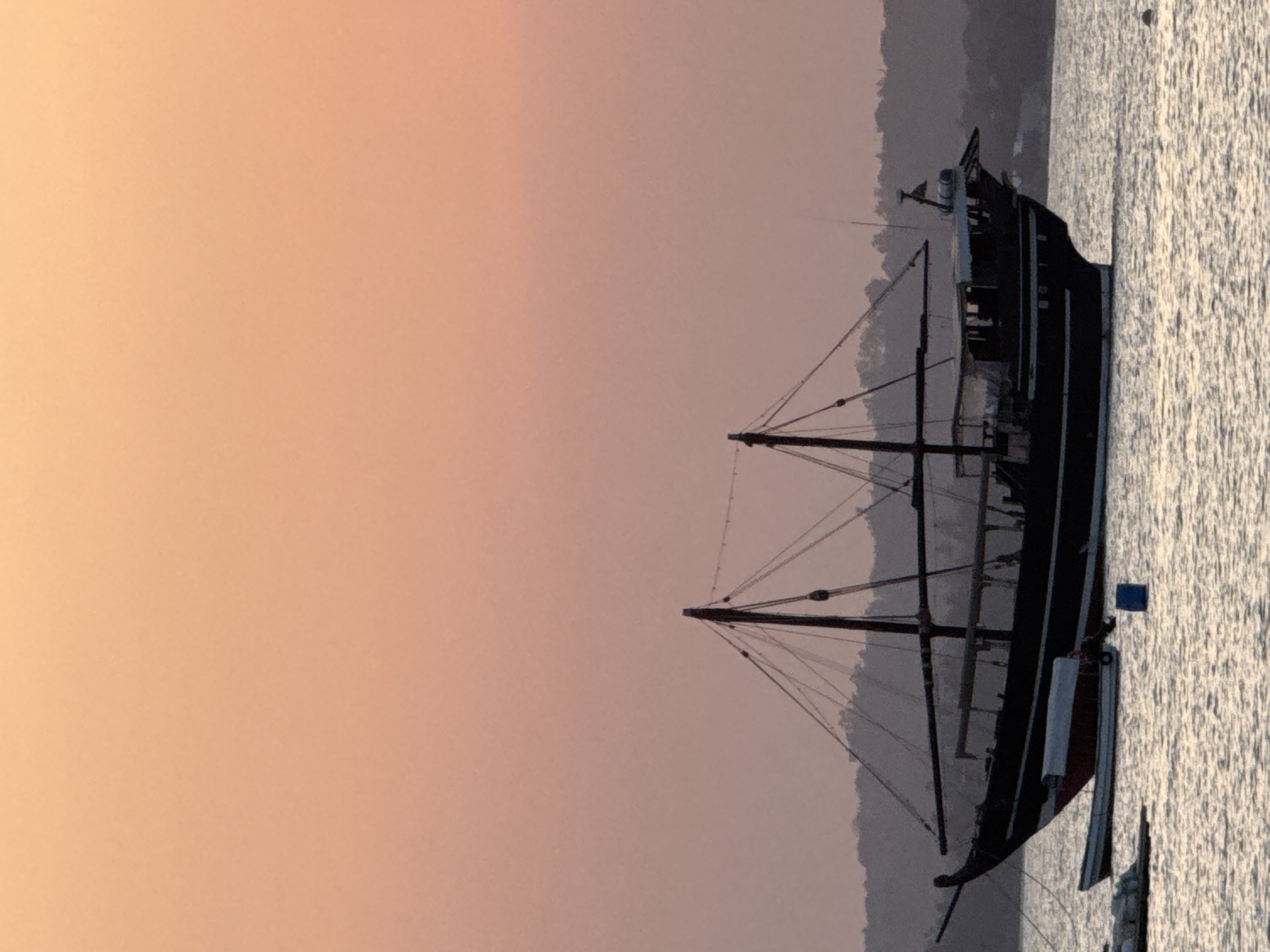 A wooden sailboat is moored in the calm waters off the coast of Dar es Salaam, Tanzania, during sunset. The sky is a gradient of soft peach and pink hues, with the warm colors reflecting on the water's surface. The sailboat, a dark silhouette against the sky, has multiple masts and rigging, suggesting it is a traditional vessel. In the foreground, the rippling water is broken by the presence of smaller boats and buoys, indicating a harbor or anchorage area. The distant shore is lined with trees, creating a hazy, dark outline against the horizon. The overall scene evokes a sense of tranquility and the end of a day.