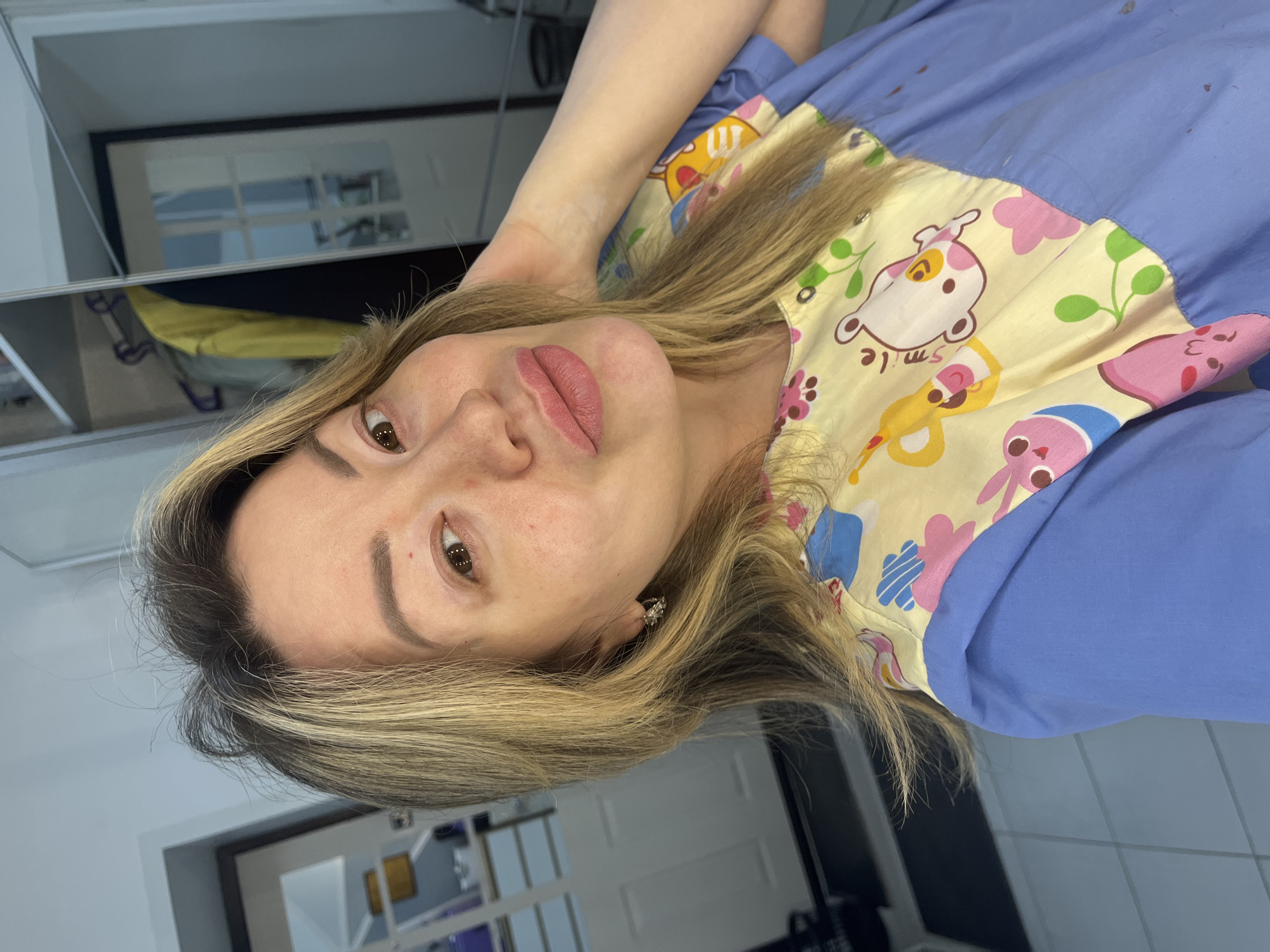 A woman with light brown hair and blonde highlights, styled in a long, straight manner, is captured in a close-up portrait, lying down and looking directly at the camera. She has full, pinkish-red lips, well-defined eyebrows, and brown eyes. She wears a light blue top with a cheerful yellow panel across the chest, featuring colorful cartoon characters including a white dog-like figure with pink spots and the word "Smile," pink bunnies, and other shapes in blue, yellow, and green. A small earring is visible in her right ear.
The background, somewhat out of focus, suggests an indoor setting with reflections on a shiny surface, possibly a mirror or glass. These reflections reveal parts of a room, including a white door with panels and a yellow object, possibly clothing or an item on a shelf or chair. The overall impression is an informal, personal photo. The provided context indicates the location is Perm, Russia.
