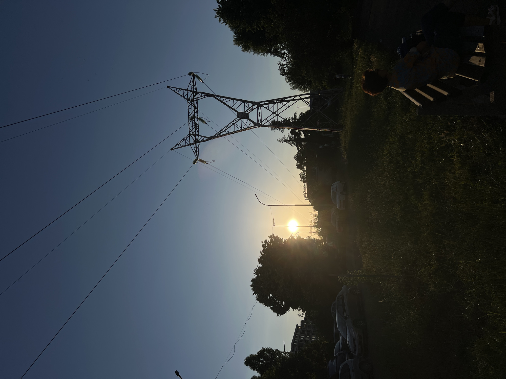 The image depicts a sunset scene near the Sea Garden in Burgas, Bulgaria, featuring a prominent electrical pylon silhouetted against a clear blue sky. The sun, low on the horizon, casts a warm glow and is partially obscured by trees on the left side of the frame, creating lens flare. Several cars are parked in a grassy area in the foreground, some with their headlights on. A person with short, reddish-brown hair is seated on a park bench in the lower right corner, facing away from the camera and towards the sunset. Their attire is indistinct due to the lighting. In the background, trees and buildings are visible, also largely silhouetted. Power lines stretch across the sky, connecting to the pylon. The overall atmosphere is serene, with the natural light of the sunset dominating the scene.