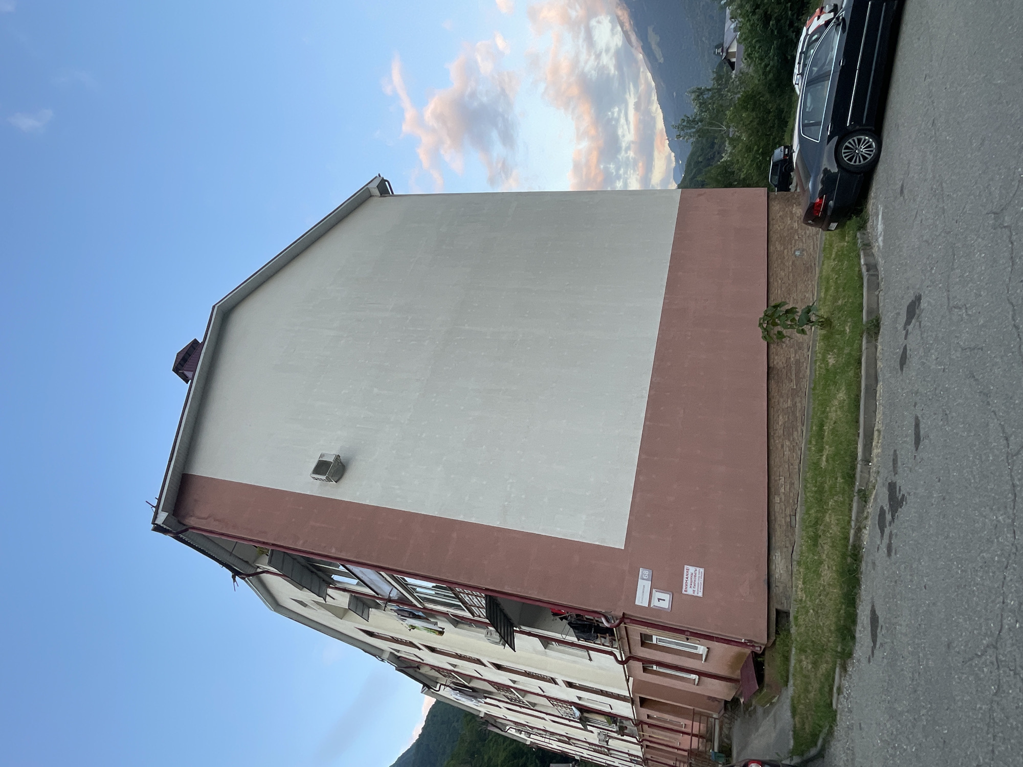 The image depicts a side view of a multi-story apartment building in Krasnaya Polyana, Russia. The building is painted in two distinct colors: the lower section and the trim around the windows and balconies are a reddish-brown, while the upper section of the facade is off-white. The building has multiple balconies with laundry hanging from them, suggesting active residency. An air conditioning unit is mounted on the off-white section of the building.
At the base of the building, there is a small patch of grass and a curb separating it from a paved road. Several cars are parked on the side of the road, including a black sedan in the foreground and two other vehicles partially visible behind it. In the background, there are lush green mountains with trees, indicating a natural, possibly mountainous, setting. The sky is a clear blue with some scattered clouds, tinged with pink and orange from what appears to be sunset or sunrise.
A sign with Cyrillic text is visible near the entrance of the building, and another smaller sign with the number "1" and "68" is also present. The architecture suggests a post-Soviet era construction. The overall scene conveys a residential area within a natural landscape.