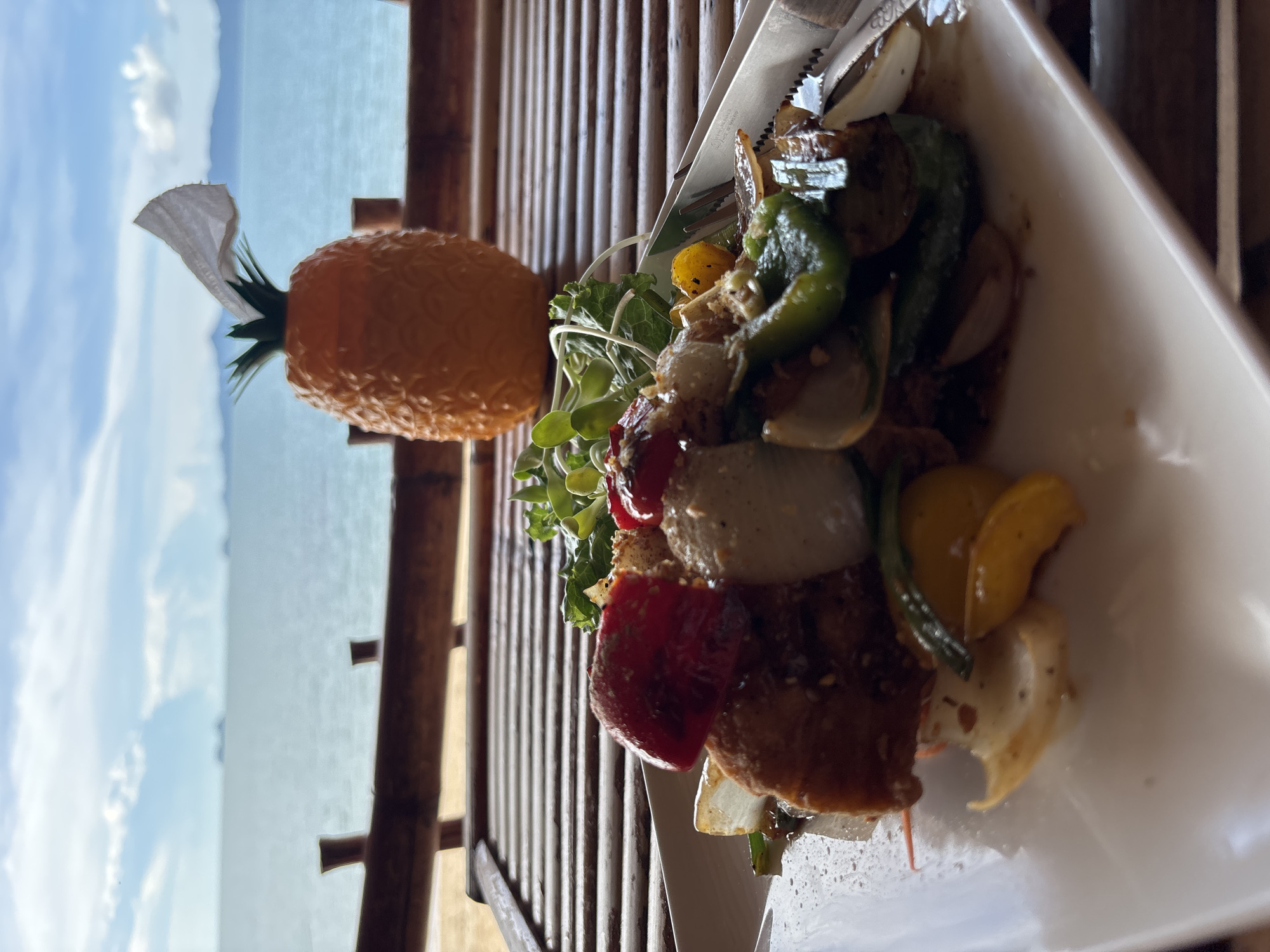 A close-up, eye-level shot captures a plate of food with a tropical beach and ocean scene in the background. The plate is white and square, with a dish containing what appears to be stir-fried vegetables and meat. Ingredients visible include chunks of red and green bell peppers, white onions, and some pieces of protein coated in a dark sauce. A small pile of fresh greens and sprouts sits atop the dish. To the right of the plate, a portion of a silver fork and knife set is visible. In the background, a decorative orange pineapple-shaped object, possibly a tissue dispenser, stands on a bamboo surface. Beyond that, the ocean stretches to the horizon under a partly cloudy sky. The overall scene suggests a meal being enjoyed outdoors, likely at a restaurant or resort with a view of the sea.