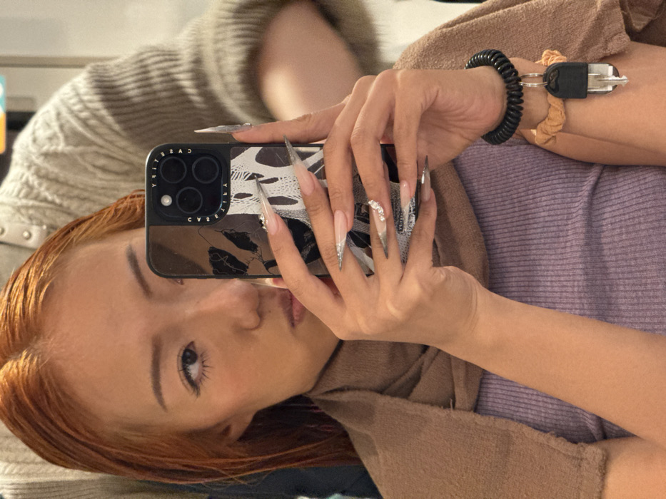 An individual with wet, reddish-orange hair is depicted indoors, likely within a hair salon in Toshima, Japan, though no specific street address or landmark is identifiable. The subject, with fair skin, occupies the central foreground, capturing a selfie using an iPhone with a black, patterned CASETiFY case. Their left eye, part of the nose, and mouth are visible, directed towards the phone screen. A brown towel is draped over their shoulders, covering a light purple ribbed top. The subject's hands exhibit long, pointed acrylic nails featuring clear designs with silver/white elements and rhinestones on the right ring finger. A black coiled wristband with a single metal key attached is visible on the right wrist, positioned above an orange scrunchie. Partially visible in the upper right background is the arm and shoulder of a second individual wearing a light-colored, textured garment, indicating the presence of another person, likely a stylist, attending to the subject's hair.