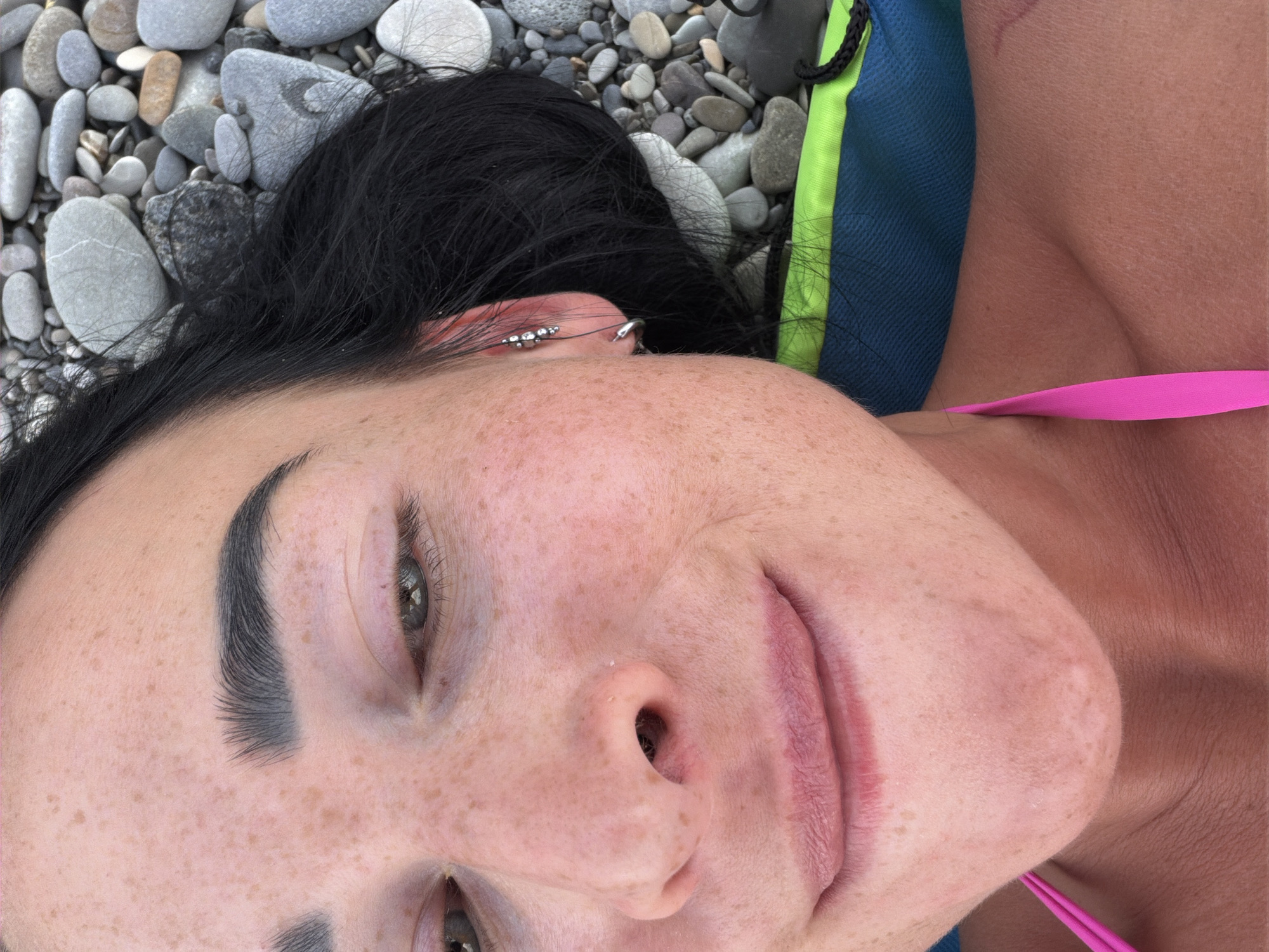 The image is a close-up, high-angle shot of a person lying on a bed of pebbles. The person has dark hair and appears to be looking upwards, with a faint smile. Their face is partially visible, showing one eye, eyebrow, nose, and lips, with freckles scattered across their skin. They are wearing a blue and green top with a bright pink strap across their chest. The background consists of various smooth, grey and white pebbles of different sizes. The lighting suggests it could be daytime, possibly outdoors given the pebbles, though no specific weather cues are evident. There is a piercing visible on the person's ear. The overall impression is one of relaxation and perhaps a candid moment captured.