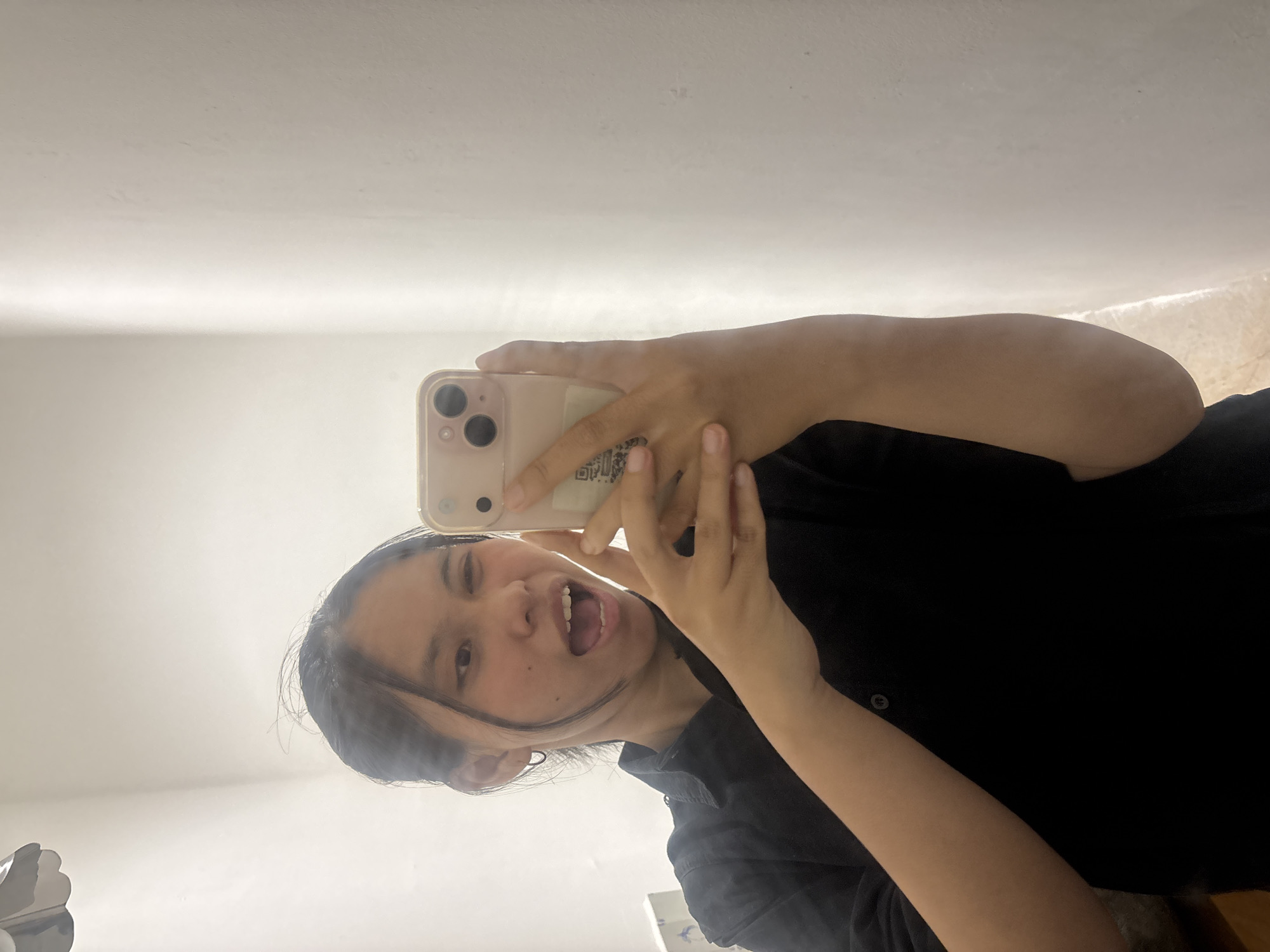 A young Asian person is taking a selfie in what appears to be a residential interior, possibly a bathroom, in South Jakarta, Indonesia. The person is wearing a black collared shirt and has dark hair pulled back. They are holding a light pink smartphone with a clear protective case in their right hand, and their left hand is partially visible supporting the phone. The person's mouth is open, and they are winking at the camera, suggesting a playful or humorous pose. The background is a plain, light-colored wall with a vertical shadow or light streak on the right side, indicating a light source. A portion of a light fixture or ceiling decoration is visible in the upper left corner.