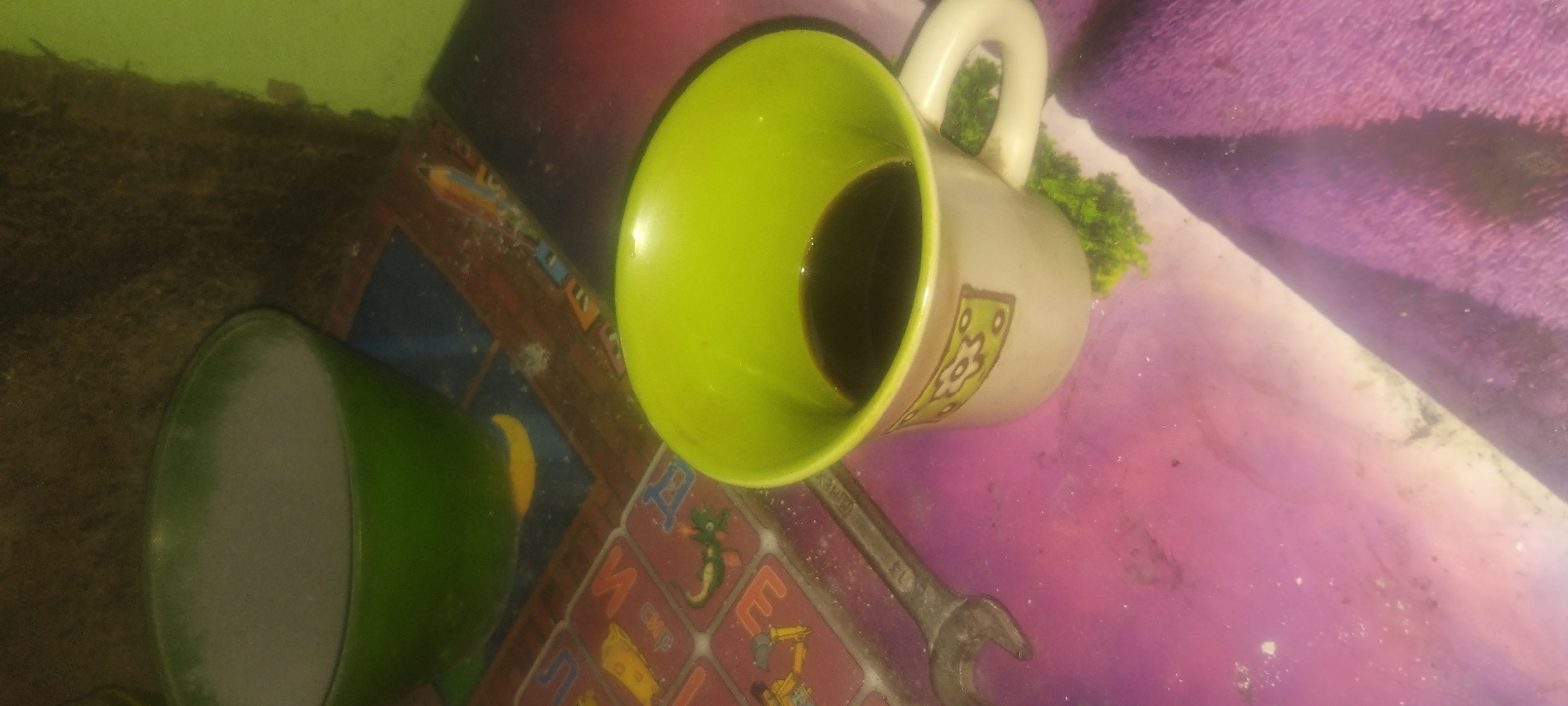 An overhead medium shot shows a wrench and a mug of coffee on a table. The mug, a beige ceramic with a light green interior and a floral pattern, is filled with dark coffee. To the left of the mug, a metal wrench rests on the table. In the background, a green bowl containing a white substance is visible. The table surface is a colorful, abstract pattern.