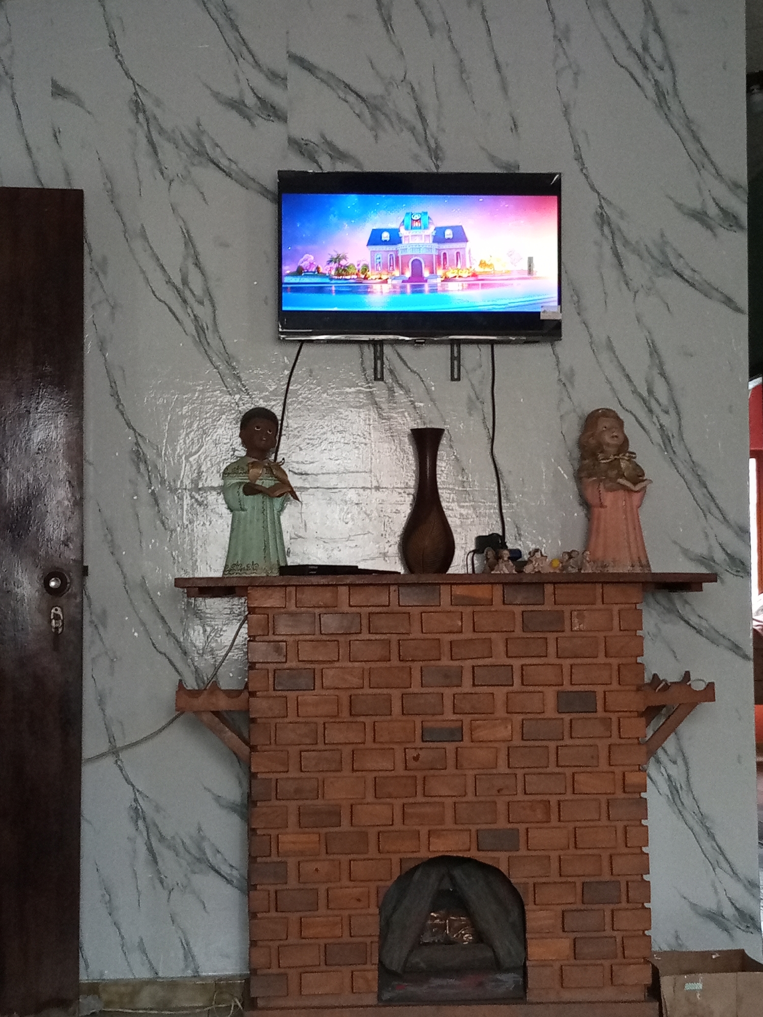The image depicts an indoor scene in Palo Negro, Venezuela. A flat-screen TV is mounted on a wall with a faux marble pattern. Below the TV is a brick structure resembling a fireplace mantel. The mantel holds a brown vase, a dark-skinned statue holding an object, a light-skinned statue in a pink dress, and various small decorative objects. The fireplace opening contains grey material. To the left of the scene is a dark-colored door with a silver knob and lock. To the right, a glimpse of what appears to be a white and red wall is visible. A cardboard box sits on the floor to the bottom right of the fireplace.