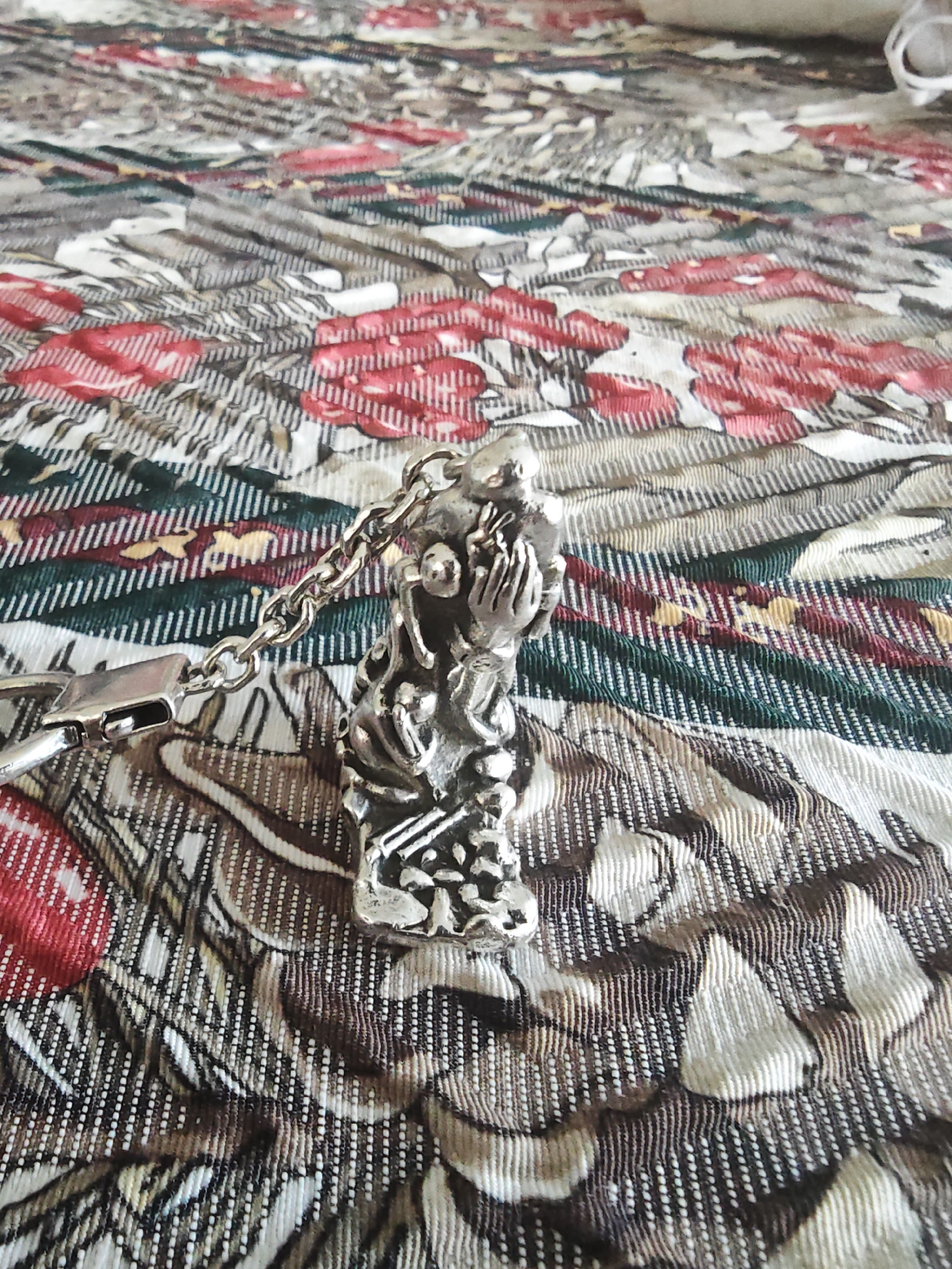 A silver colored keychain pendant in the shape of a kneeling human figure with hands clasped is shown from a high angle. The pendant is attached to a silver colored chain and a clasp. The pendant and chain are resting on a patterned fabric surface with red and green floral designs. The image was taken indoors on January 18, 2023, in Barntrup, Germany.