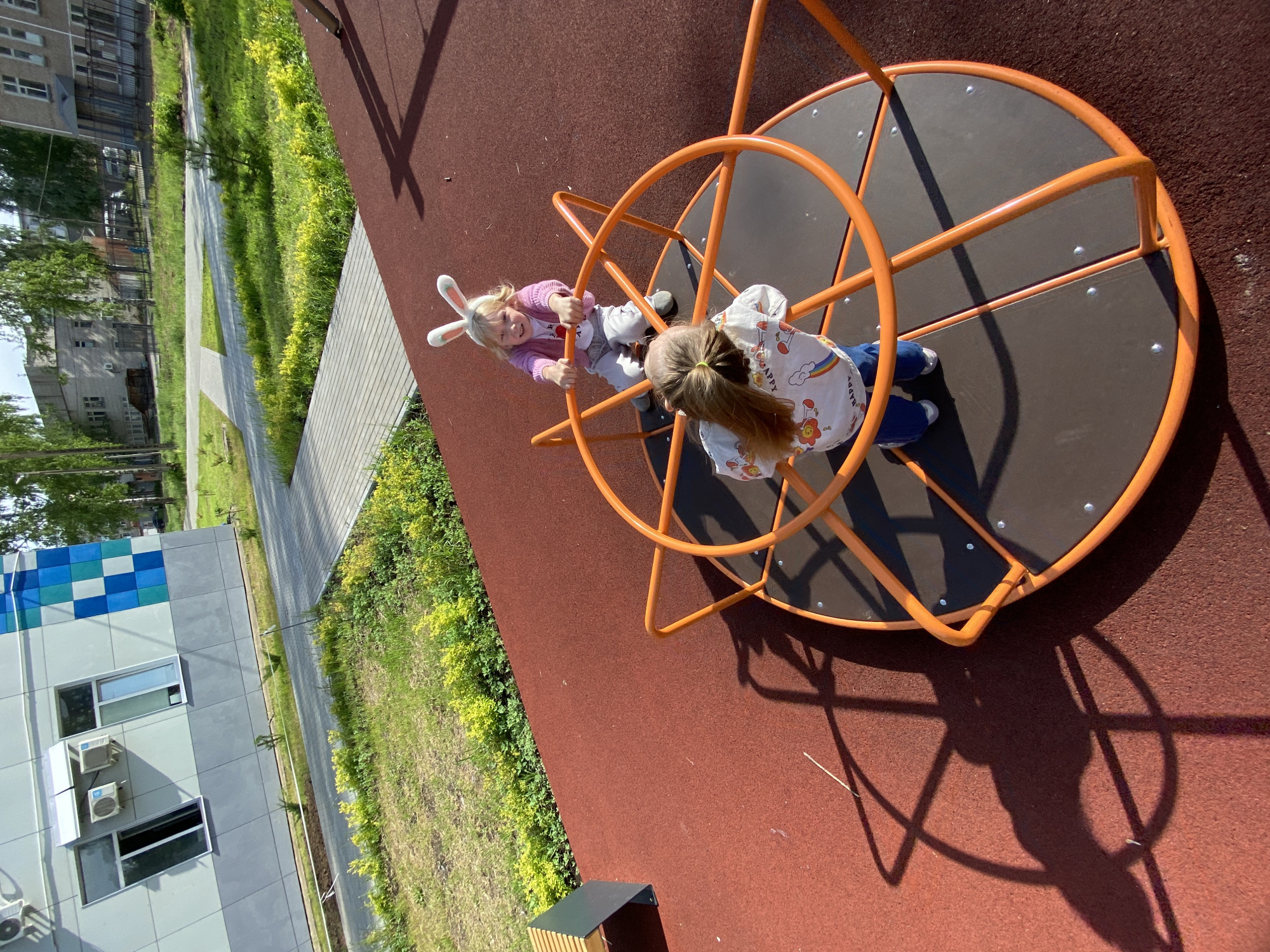 Two young girls are on a roundabout at a playground. One girl is wearing bunny ears, a pink cardigan, and grey pants. The other girl has brown hair tied back with a yellow hair tie and wears a white shirt with a rainbow and flower design and blue jeans. The roundabout is orange and black. The playground surface is a reddish-brown color. The playground is surrounded by green grass and bushes. In the background, there is a building with blue and white tiled sections and windows with air conditioning units. The scene is located in the city of Chaikovsky, Russia.