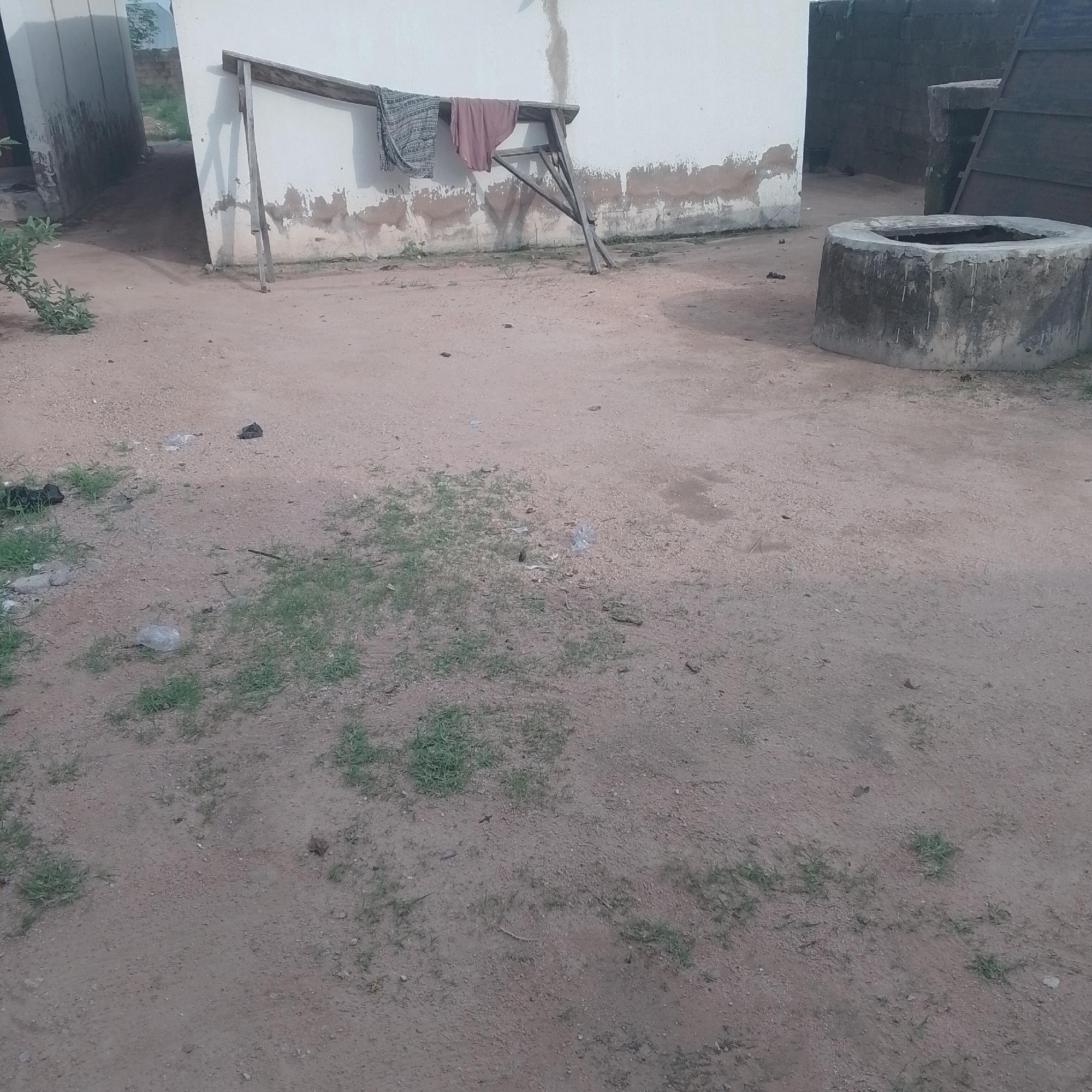 The image depicts an outdoor scene in Jalingo, Nigeria, characterized by a dirt ground with scattered patches of grass. In the background, a portion of a building with a white, weathered wall is visible. Attached to the wall is a makeshift wooden structure holding two pieces of fabric that appear to be clothes, possibly drying. To the right of the building, a circular, concrete structure, likely a well, is present. The well has a weathered appearance with streaks of discoloration on its exterior. The overall impression is that of a simple, rural or semi-urban setting.