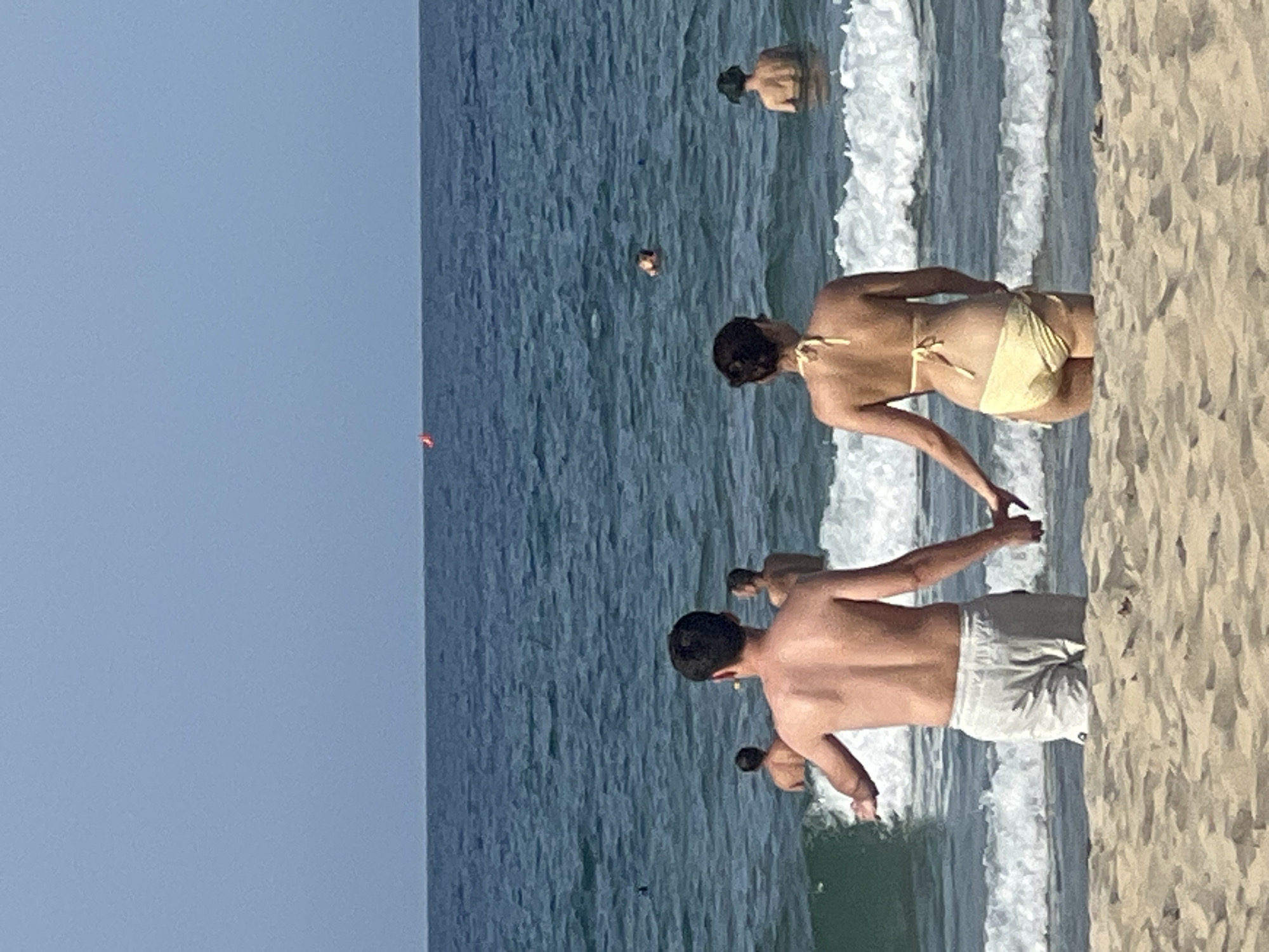 A couple is holding hands at the edge of the ocean on a beach, with the man facing away from the camera wearing light-colored swim trunks and the woman facing away from the camera wearing a yellow bikini. They are standing in shallow water with waves lapping at their legs. In the background, other people are visible in the ocean, some swimming and others standing. A bright orange buoy is floating on the horizon. The sky is clear and blue. The sand in the foreground is light brown and textured. The image appears to be taken on a sunny day in Da Nang, Vietnam.