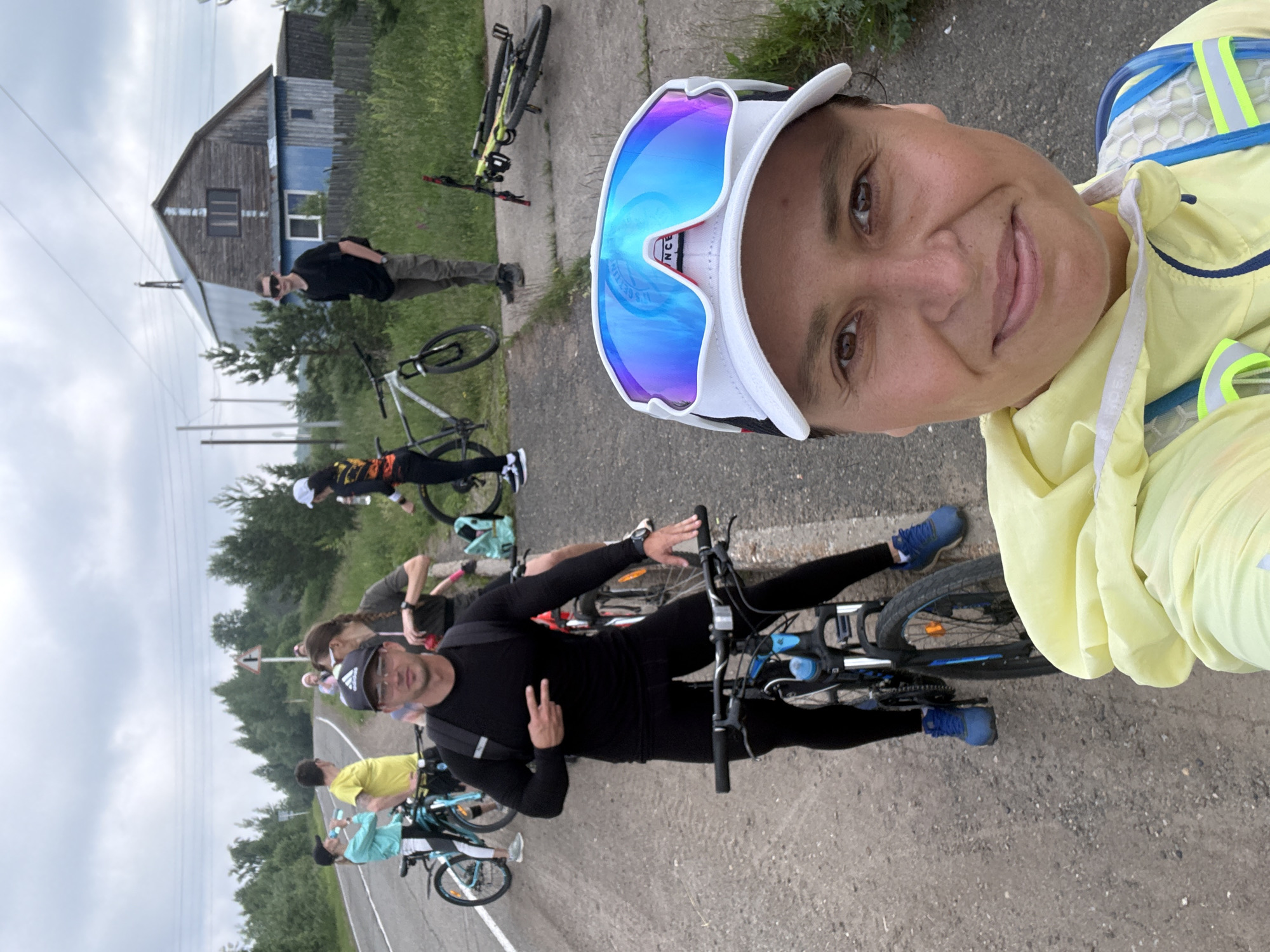 A group of people are gathered on the side of a rural road in Kharnavy, Russia. The foreground is dominated by a woman wearing a yellow jacket and a white visor with reflective blue lenses. She is taking a selfie, her face partially visible. Behind her, several individuals are with their bicycles. One man in black sportswear is posing with two fingers up, standing next to his bike. Other people, some with bikes, are further in the background. A rustic wooden building with a metal roof and a blue lower section is visible behind the group, situated amidst greenery and trees. The sky is overcast. The road surface appears to be asphalt or packed gravel.