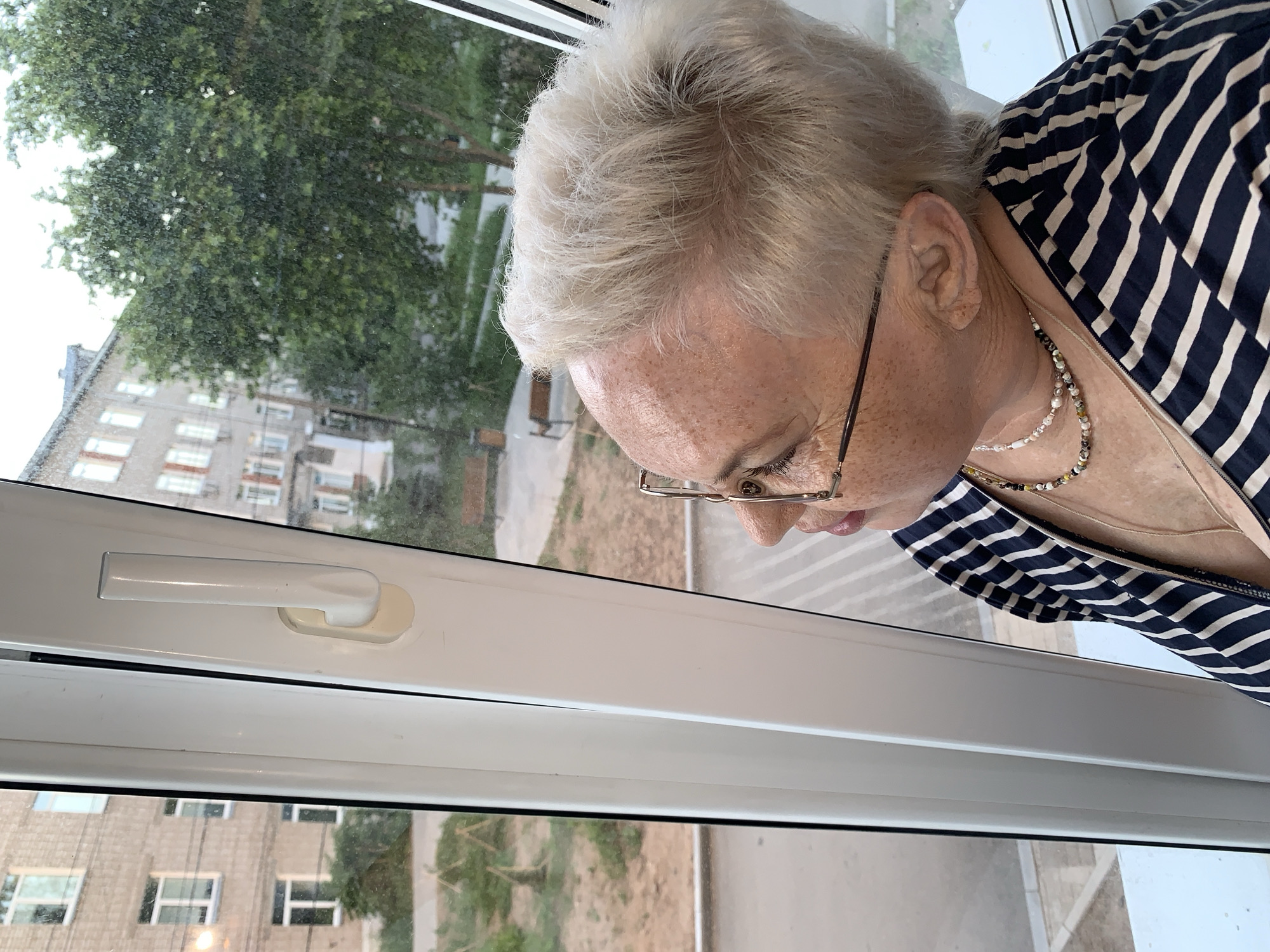 A medium-range shot depicts a fair-skinned, white-haired, freckled woman wearing glasses and a navy and white striped shirt, peering out of a window. She is wearing at least two necklaces. The location of the photo is Chaikovsky, Russia. The window features a white handle. Through the window, a residential building and trees are visible.