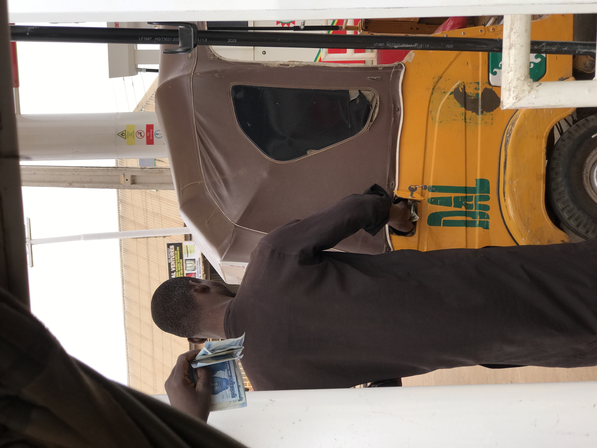 A man in a brown shirt is paying for something while holding a wad of naira notes. He is standing next to a yellow tricycle with a brown canopy. The tricycle has green writing on it and a tire is visible. A sign for "AL VENTURES" is visible in the background. There is an indication of Abuja, Nigeria.