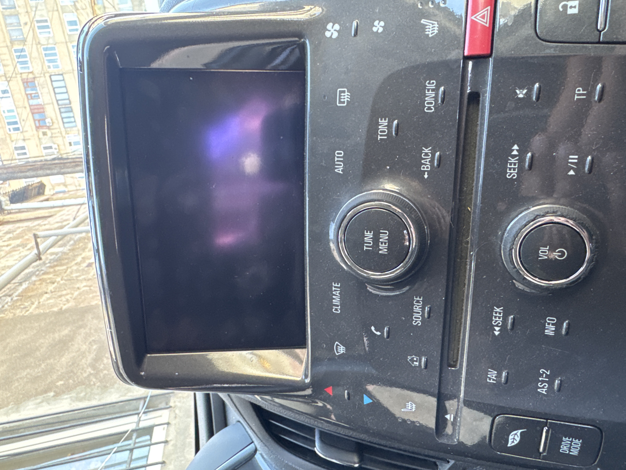 The image shows the dashboard of a vehicle, likely a car. The dashboard features a touchscreen display, surrounded by a glossy black frame. To the right of the screen is a control panel with various buttons and knobs, including climate control options, audio controls (such as tune/menu, volume), and drive mode settings. The interior appears somewhat dusty. The background visible through the windshield shows buildings, suggesting an urban environment.
There is no direct indication to suggest this photograph was taken in Yerevan, Armenia.