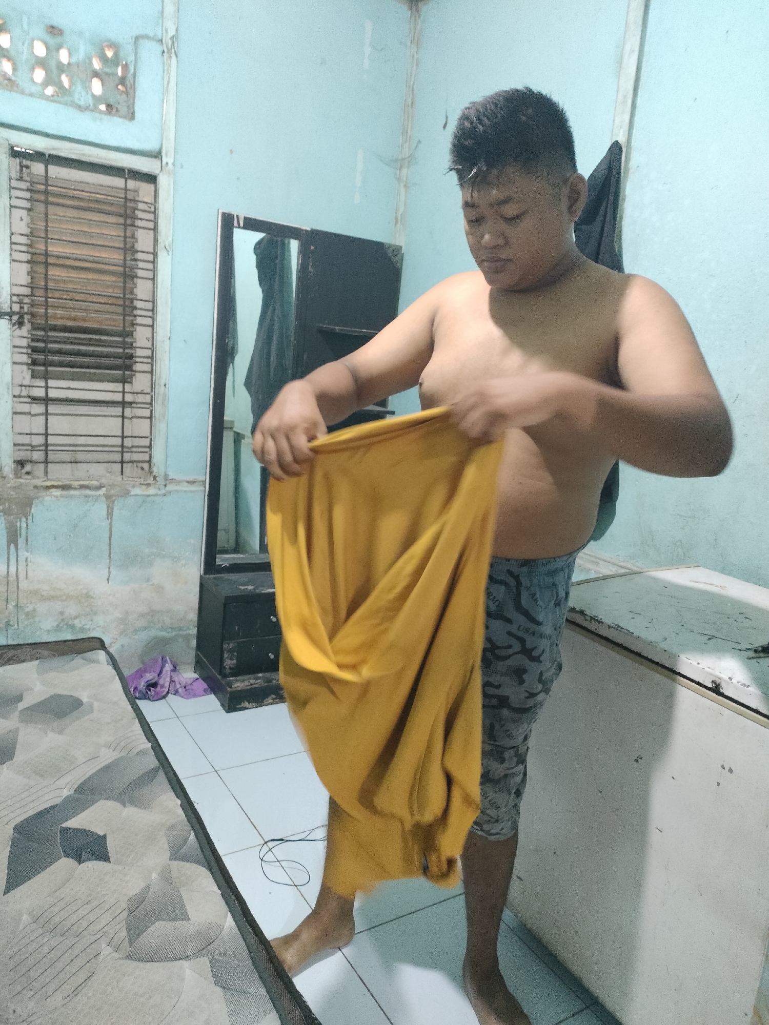 In a sparsely furnished room, a shirtless young person with dark, spiky hair is folding a bright yellow garment. The person is wearing gray camouflage shorts. The room's walls are painted a light blue and show signs of wear, with streaks of dirt and a partially open window with metal bars. A mattress with a geometric pattern is visible on the left, and a black dresser with a mirror stands in the background. A white refrigerator or similar appliance is positioned to the right. The floor is tiled in a light color. The overall impression is of a simple living space, possibly in a residential area of Pekanbaru, Indonesia, where the image was captured.