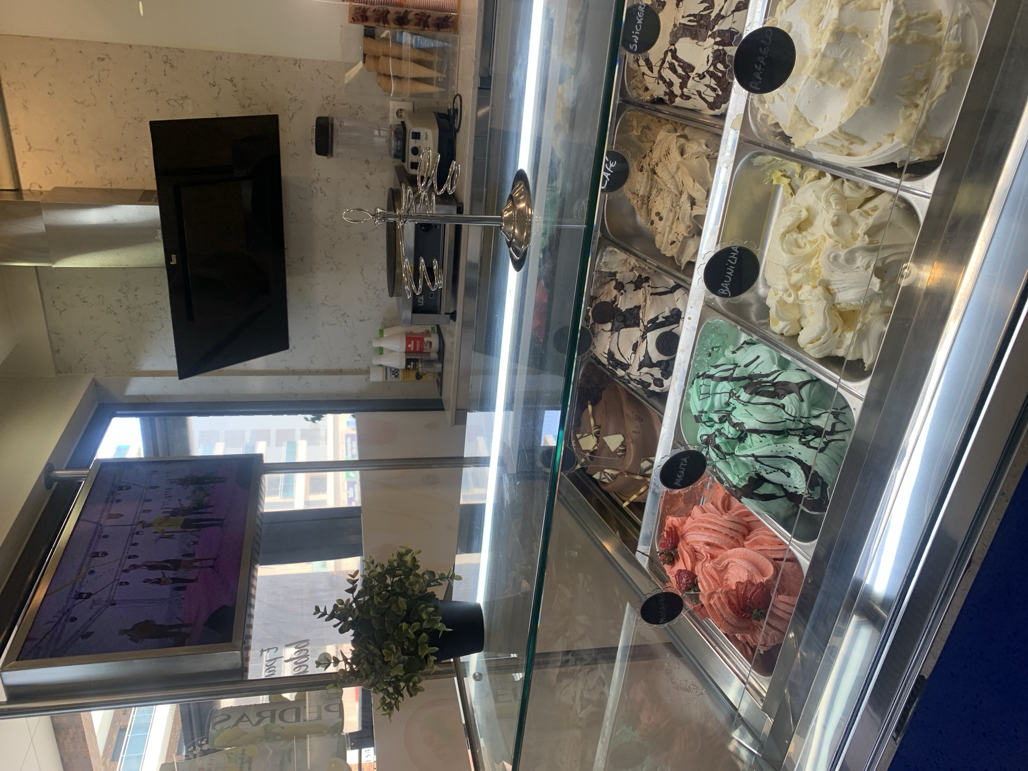 An indoor shot captures an ice cream display case filled with various flavors, including strawberry, mint, chocolate, vanilla, and what appears to be a coffee or caramel flavor, each labeled with small black signs. Above the display, a silver rack holds ice cream cones. In the background, a commercial blender and what looks like a crepe maker are visible on a counter. A flat-screen TV displays a group of people on a stage, possibly a performance. The scene suggests a dessert shop or cafe.