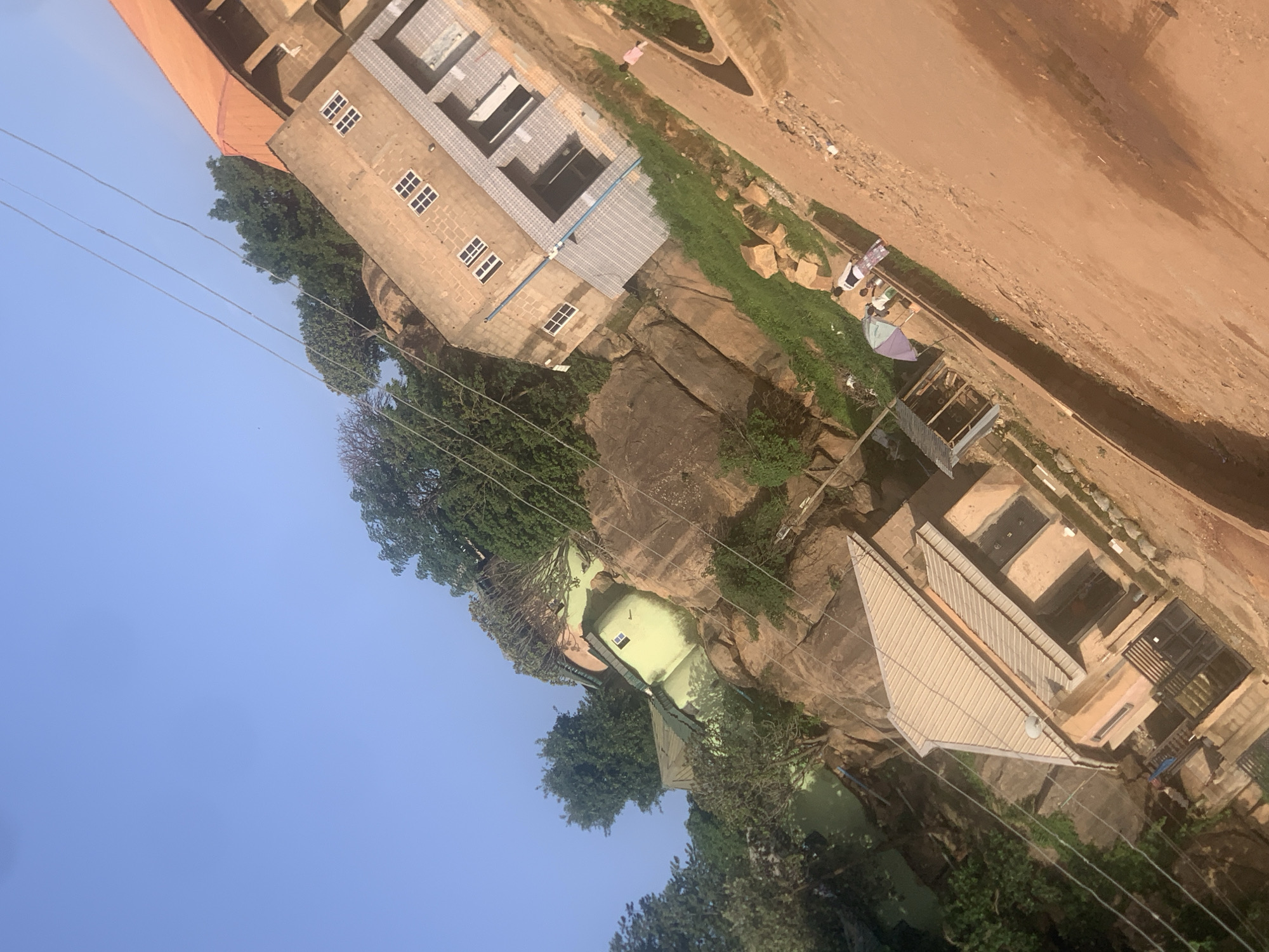 The image depicts an aerial view of a multi-level building complex situated on a rocky, uneven terrain. The architecture is varied, with some sections featuring tiled roofs and others with corrugated metal roofing. One building has a distinctive orange-tiled roof and a facade constructed from light-colored bricks with multiple small windows. Another building has a pale green exterior. Several large rocks are visible amidst dense green foliage.
In the upper right portion of the image, a dirt path runs alongside the buildings, with a small group of people and some structures suggestive of market stalls. There is a purple and white umbrella visible near these stalls. The environment appears to be a rural or semi-urban setting. The clear blue sky suggests fair weather, and the lighting indicates it is daytime, likely mid-morning or afternoon, due to the shadows cast. The overall impression is one of a developing area, with ongoing construction or development visible. No visible text is present in the image.