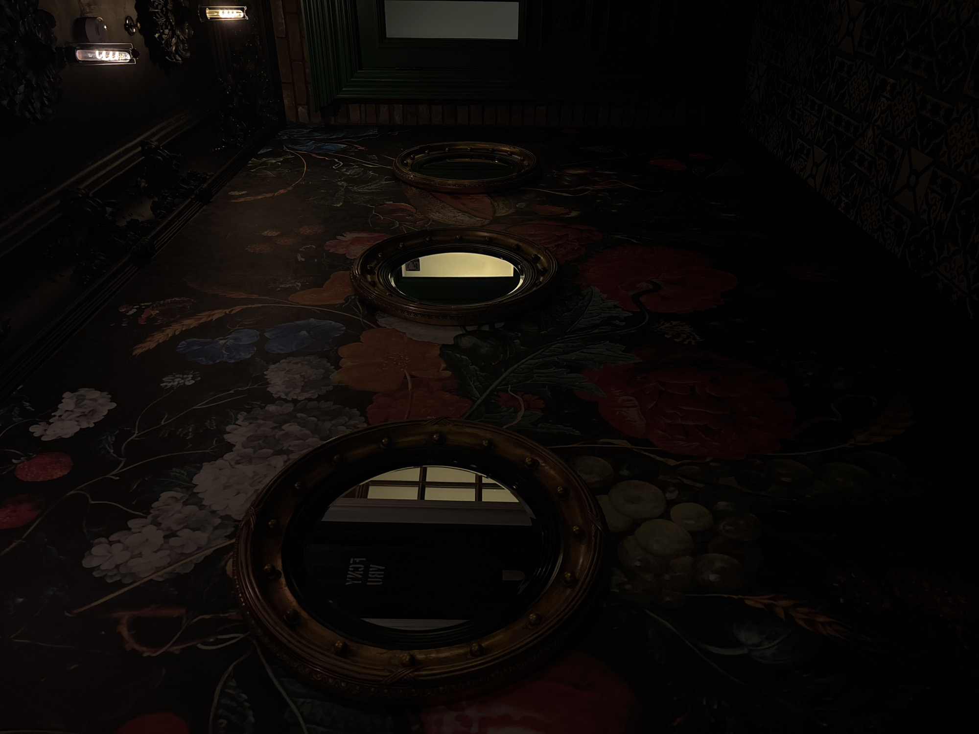 The image depicts a dimly lit interior space with three circular mirrors placed at different levels along a wall covered with a dark floral wallpaper. The mirrors reflect a brightly lit area, possibly a hallway or room with light-colored walls and what appears to be windows with blinds or bars. The wallpaper features large flowers in shades of red, white, blue, and orange against a black background. The mirrors have ornate, gold-colored frames. A patterned wall or fabric is visible on the right side of the image, adding to the overall richness and complexity of the space. The image was captured in Saint Petersburg, Russia.