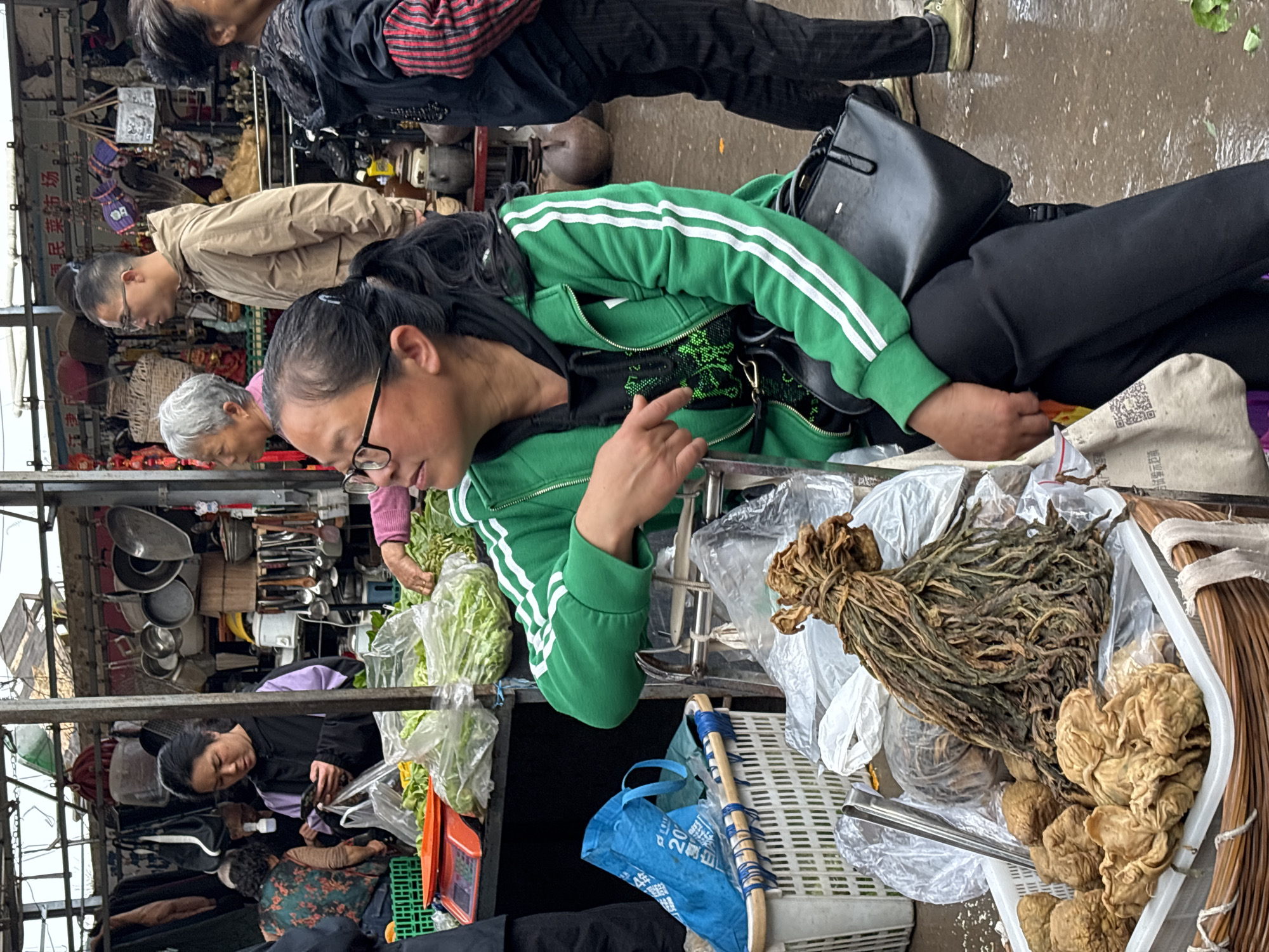 The image captures a vibrant street scene in Jiangjin, China, likely a bustling market. Several individuals are visible, including a woman in a green tracksuit jacket pointing her finger, a seated woman in a black jacket focused on something below her, and other people browsing and interacting with vendors. The environment suggests an outdoor marketplace with metal stall structures visible. The stalls are stocked with goods like leafy greens, dried vegetables, mushrooms, and household items such as pots, pans, and kitchen utensils. Additional details include blue bags, wicker baskets, and plastic packaging materials for the various products, adding to the sense of a busy and authentic marketplace in Jiangjin, China.