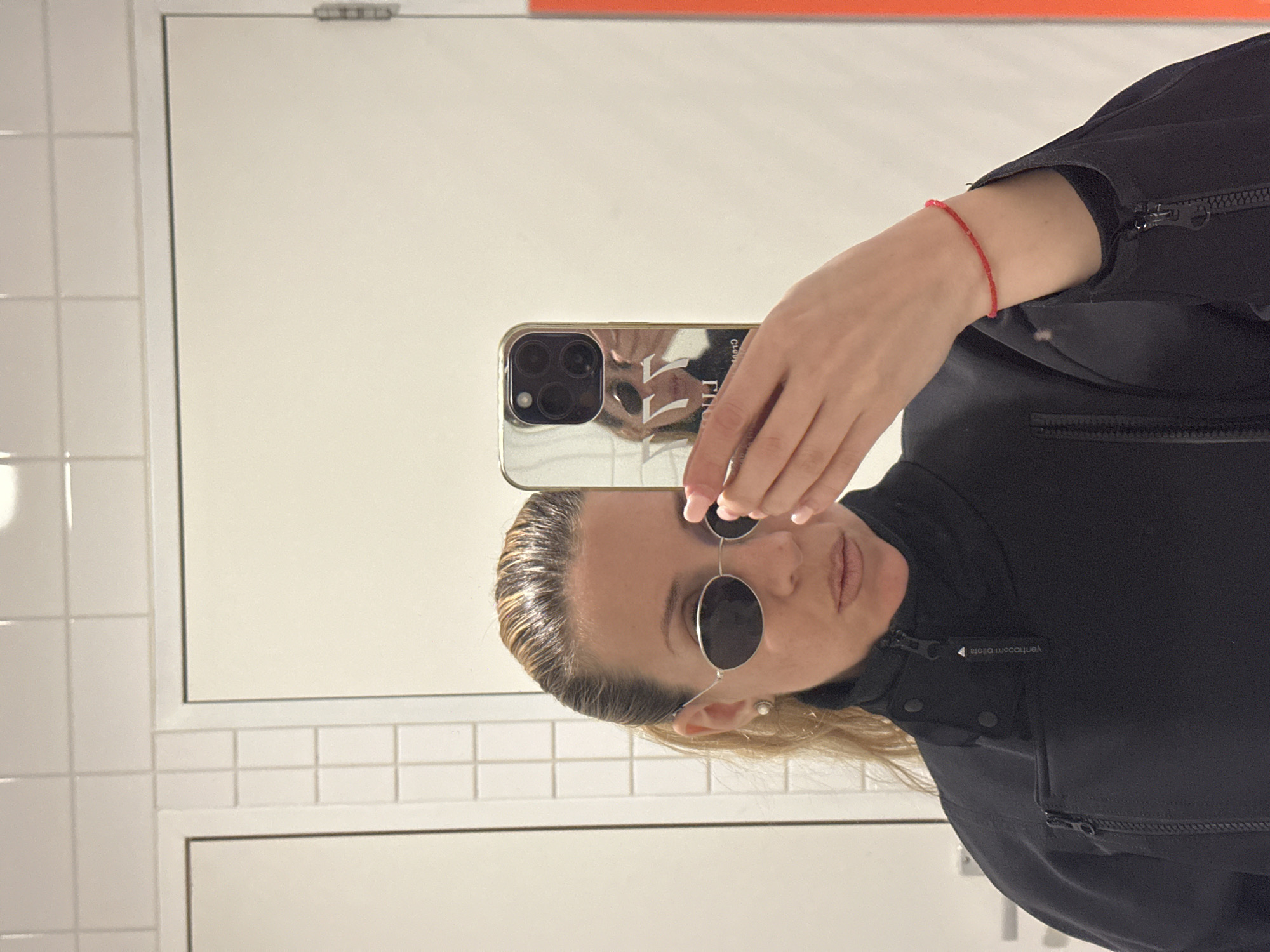 The image depicts a person in a bathroom, likely a public restroom. The person is holding a silver smartphone with a triple-lens camera, taking a selfie in a mirror. They are wearing round, dark sunglasses, a black jacket with zippers and buttons, and a thin red bracelet on their left wrist. Their hair appears to be blonde and styled. The background consists of white tiled walls. No definitive evidence indicates the location as Amsterdam.