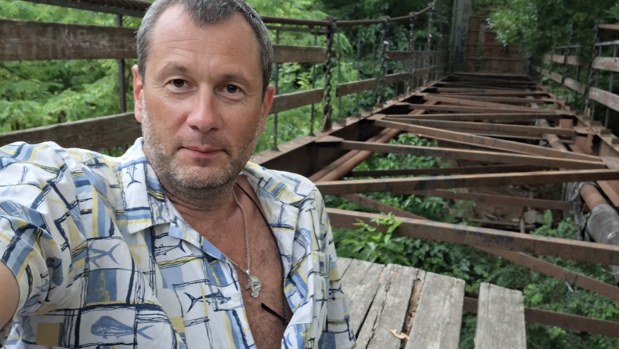 A middle-aged man with short graying hair and a beard is shown from the chest up, taking a selfie on a dilapidated wooden and metal bridge. He is wearing a short-sleeved, collared shirt with a pattern of blue, yellow, and white squares, some containing fish motifs. A silver chain with a pendant is visible around his neck, resting on his chest, which is partially visible through the open buttons of his shirt.
The bridge appears to be in disrepair, with missing wooden planks and rusted metal supports. It is surrounded by lush green foliage, suggesting an outdoor, possibly natural, setting. The background is dominated by the bridge structure and greenery. The man is looking directly at the camera with a slight smile. The overall impression is a candid moment captured while exploring an old structure.
The prompt requires me to identify the location in Chișinău, Moldova. However, the image itself does not contain any discernible landmarks, signs, or architectural features that would allow for specific identification of Chișinău, Moldova, or any other specific city. Therefore, I cannot provide details about its location.