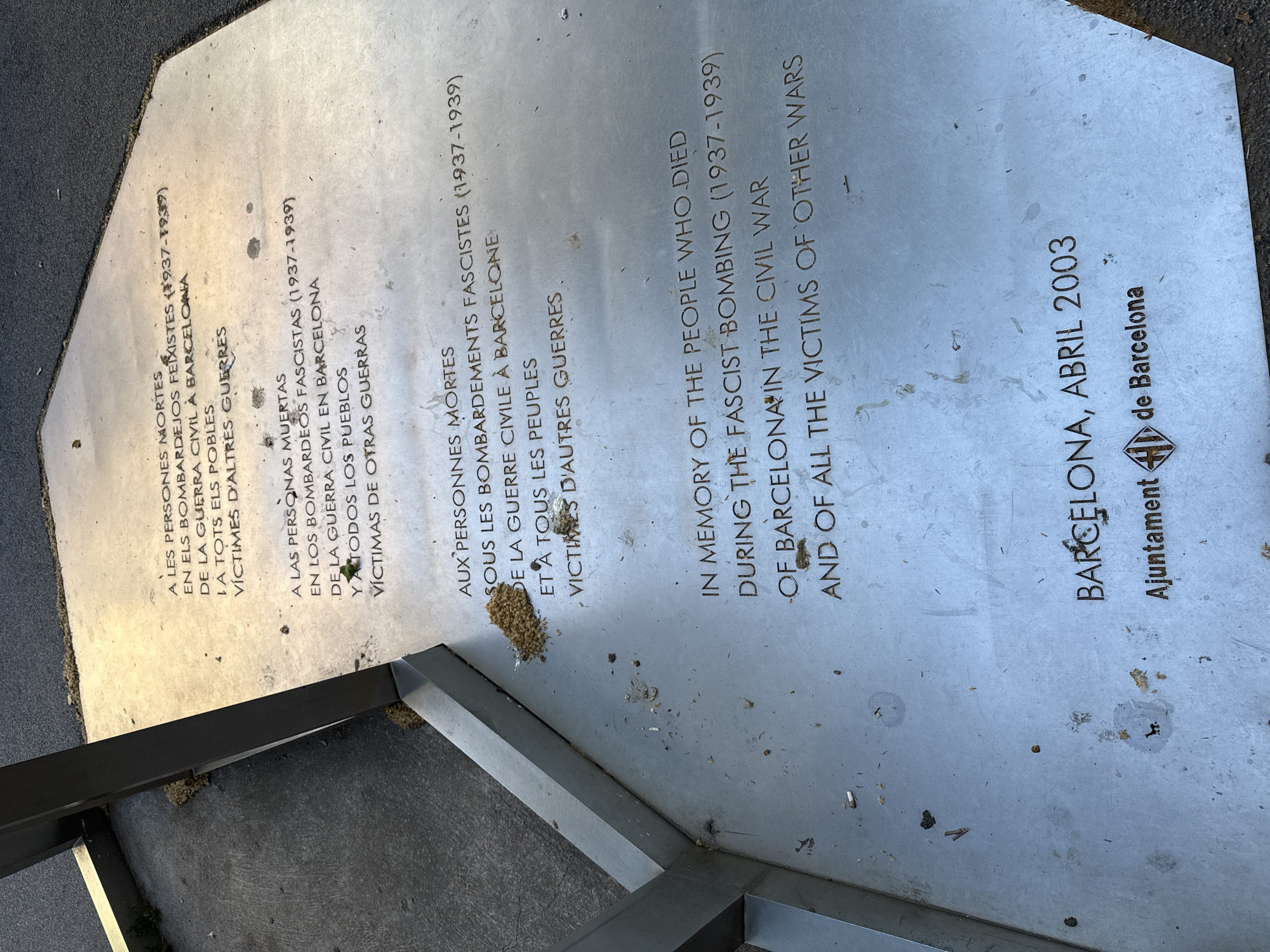This is a close-up shot of a commemorative plaque located in Barcelona, Spain. The plaque is made of a metallic material, likely stainless steel, and is inlaid into the ground. The text is engraved and filled with a dark material.
The plaque commemorates the people who died during the fascist bombing of Barcelona during the Spanish Civil War (1937-1939), as well as all the victims of other wars. The text is written in Catalan, Spanish, French, and English.
At the bottom of the plaque, it reads "BARCELONA, ABRIL 2003" followed by the logo of "Ajuntament de Barcelona" (Barcelona City Council). The plaque is surrounded by a metal frame. There is debris scattered on the plaque, including organic material like seeds. The photograph is taken outdoors during daylight.