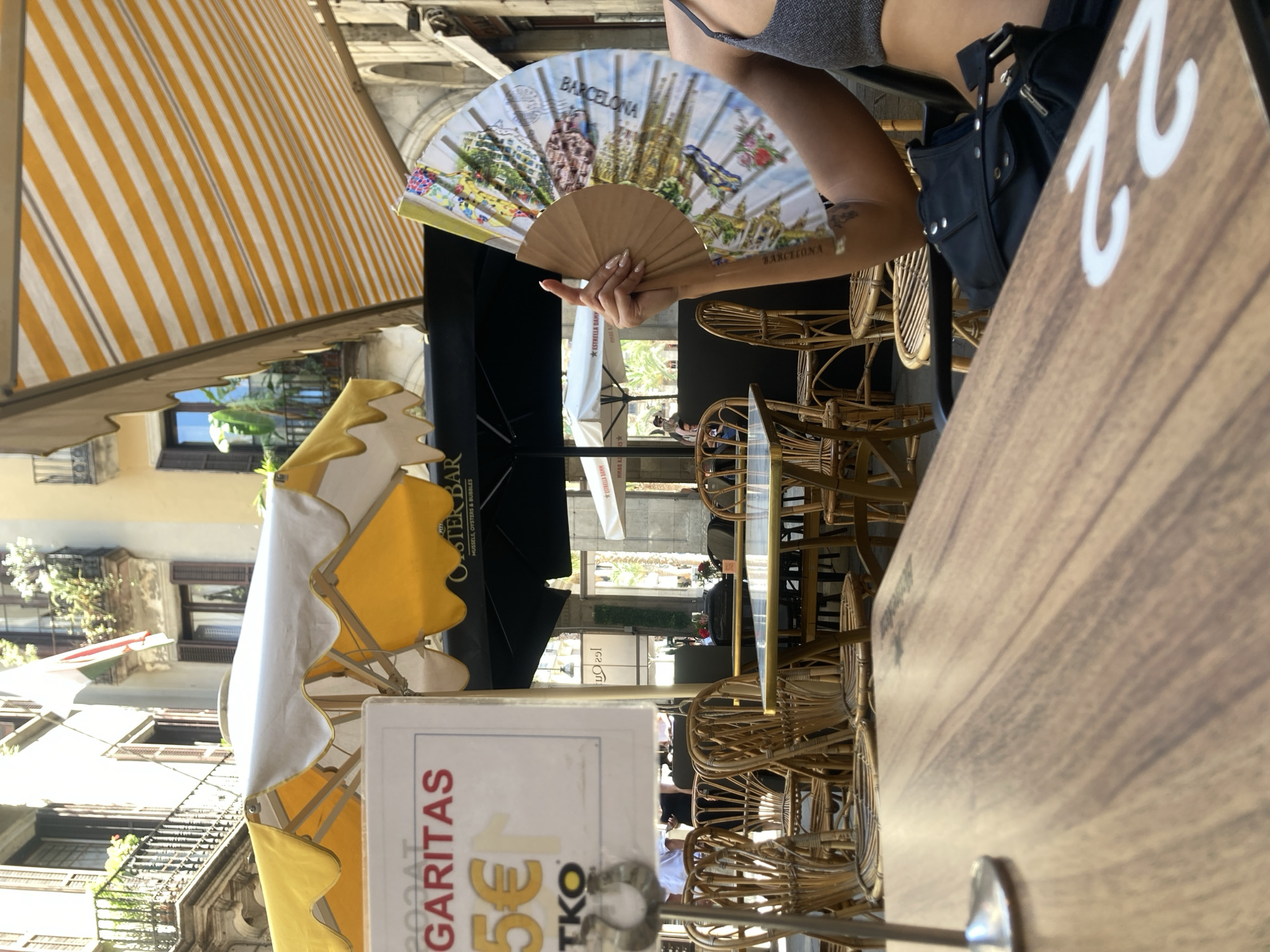 The image depicts an outdoor cafe scene on a sunny day in Barcelona, Spain. A person is partially visible on the right, holding an open fan decorated with images of Barcelona landmarks. In the foreground, a wooden table with the number '22' painted on it is visible. Behind the table, several empty rattan chairs are arranged around tables under yellow and white striped awnings. A sign with "TKO" and "5€" is attached to a metal stand. In the background, buildings with balconies and flags are visible, suggesting a European city setting. The overall atmosphere is relaxed and typical of a cafe during daytime.