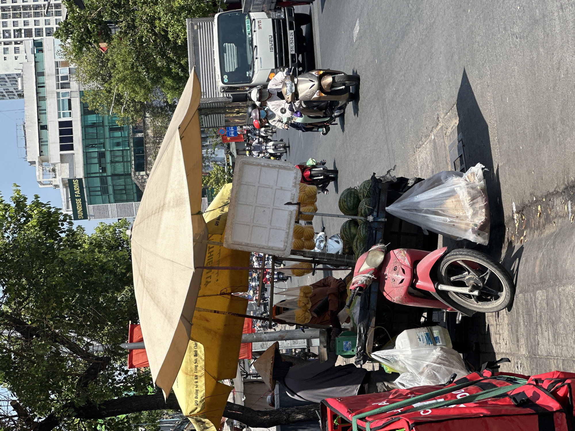 The image depicts a street scene in Ho Chi Minh City, Vietnam. A vendor is selling produce, likely fruit, from a stall shaded by a large, light-yellow umbrella. The stall is positioned at the edge of the sidewalk, with a red motorcycle parked nearby. The vendor is wearing a traditional conical hat. The street behind the stall shows a moderate flow of traffic, including motorcycles, cars, and a truck. The "SUNRISE FARMS" building is visible in the background. Signage on the umbrella reads "120 Đường số 9, Khu Trung Sơn, Bình Chánh," indicating the address.
