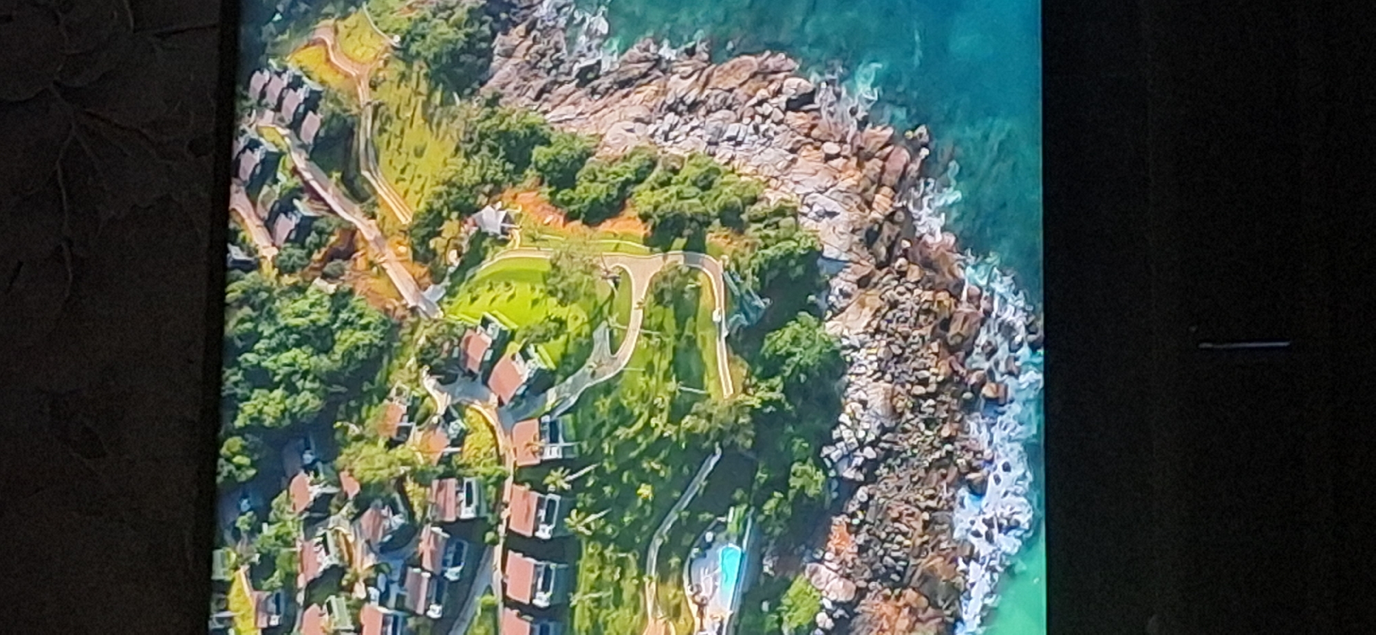 The image depicts an aerial view of a coastal resort area with several modern villas scattered across a lush green hillside. The villas, characterized by their red-tiled roofs and white walls, are interconnected by winding pathways. The hillside is densely vegetated with trees, and some areas are manicured into lawns. In the foreground, rocky shorelines meet the turquoise ocean, with waves crashing against the rocks. A swimming pool is visible near one of the villas. The scene suggests a luxurious and serene vacation destination. This image is not from Bila Tserkva, Ukraine, as Bila Tserkva is an inland city and not located on the coast.