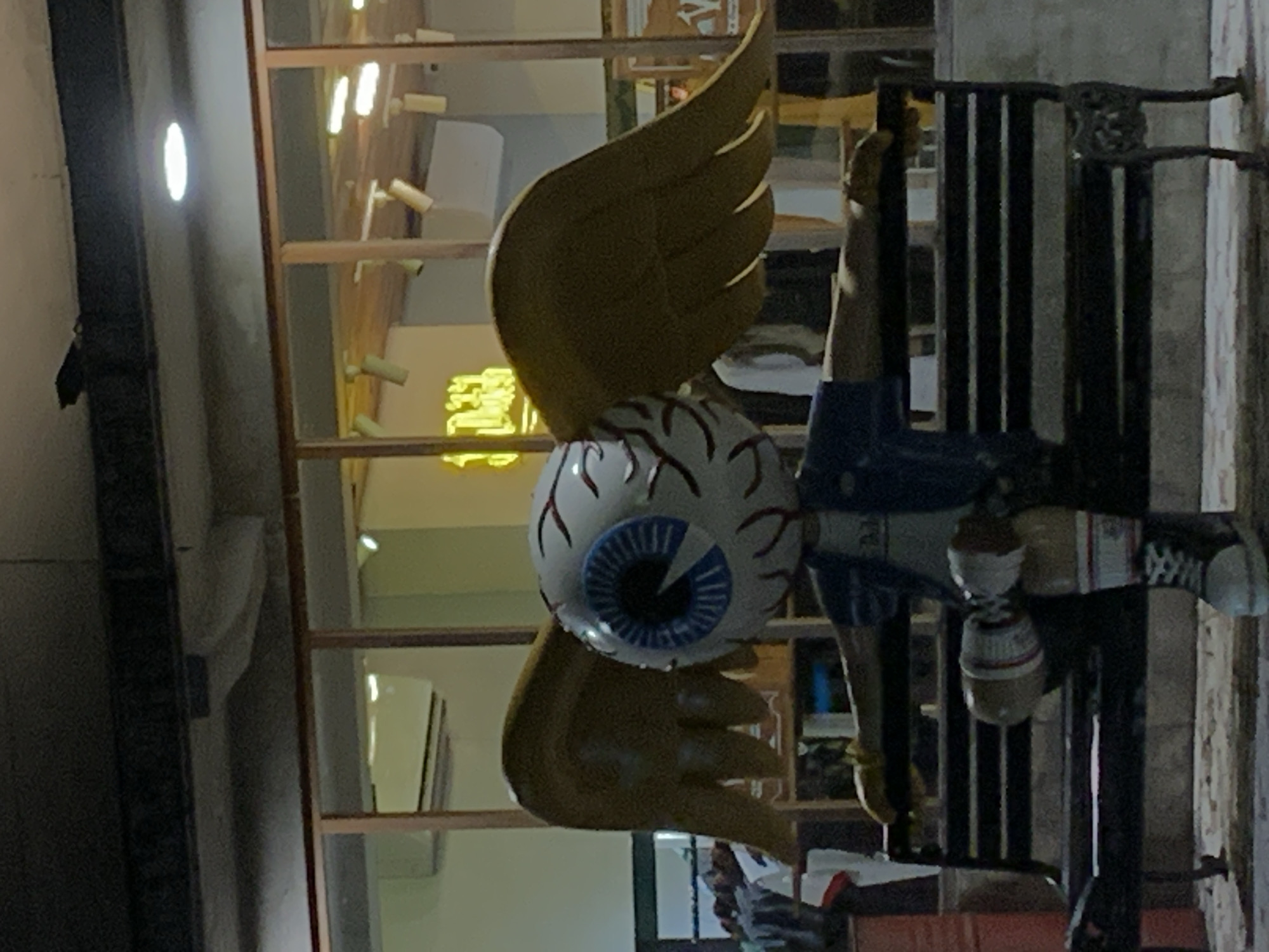 The image shows an outdoor scene, possibly in Ubud District, Indonesia, featuring a unique art installation. This installation is composed of a large, stylized eyeball with golden wings attached, and a person in a blue denim jacket and black shoes mounted on top of a metal bar. The artwork is positioned outside a building with a glass window front, where an interior is visible with shelves and other items. The light suggests that the picture was taken during the evening or nighttime. The statue appears to be a kind of mannequin, with two cans/bottles positioned where the neck would be. There appears to be a second person behind the installation, lower to the ground, but that person cannot be made out in any detail.