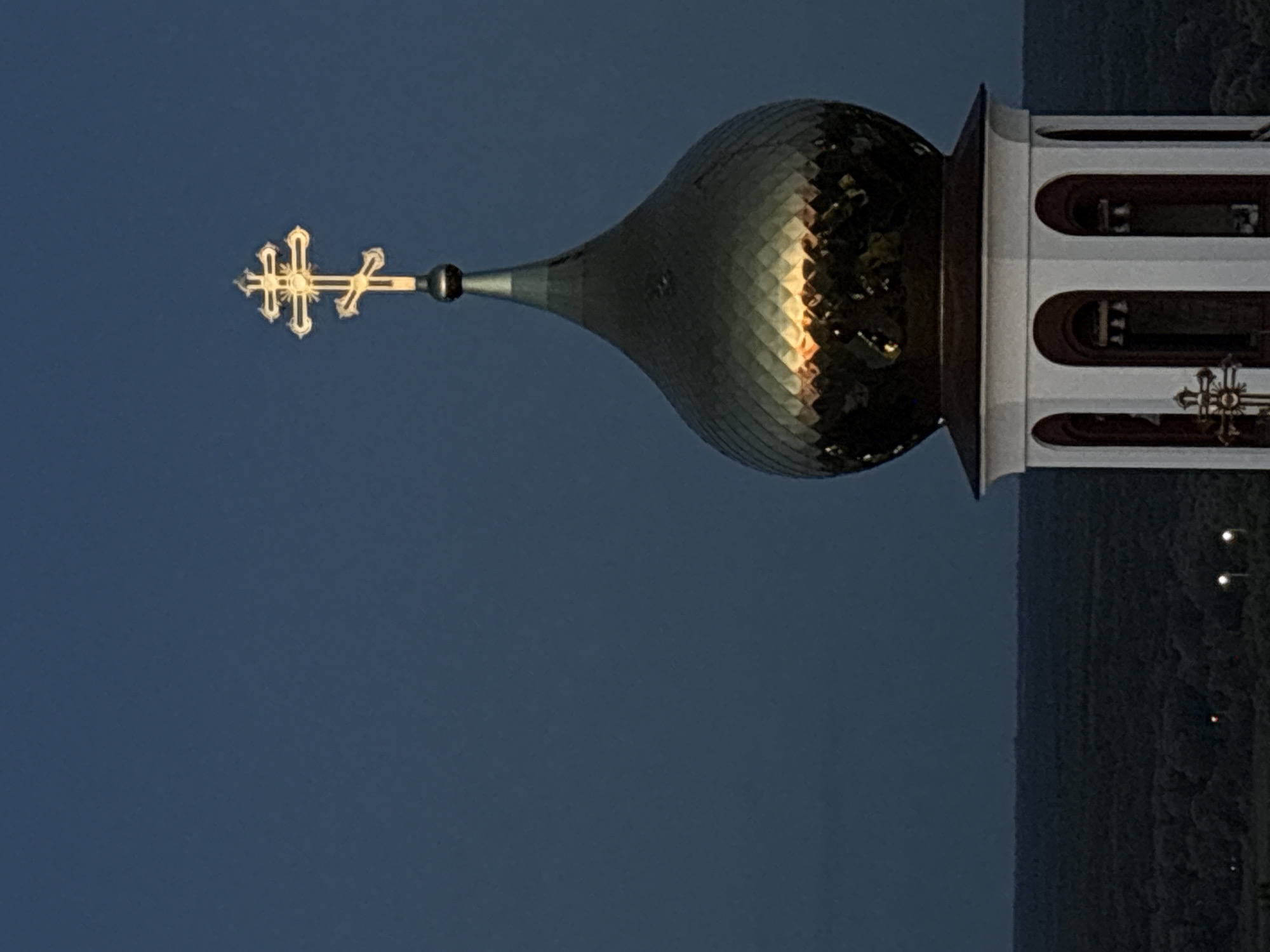The image shows the golden dome and cross of an Orthodox church in Bobrov, Russia, set against a dark blue twilight sky. The dome is textured with diamond-shaped panels that reflect the warm glow of the setting sun or artificial lights. Below the dome, a white bell tower with arched openings and visible bells can be seen. A smaller, ornate cross is visible on the lower part of the bell tower. In the background, a dark, tree-covered landscape stretches to the horizon, with a few scattered lights visible in the distance. The overall scene evokes a sense of tranquility and religious solemnity.