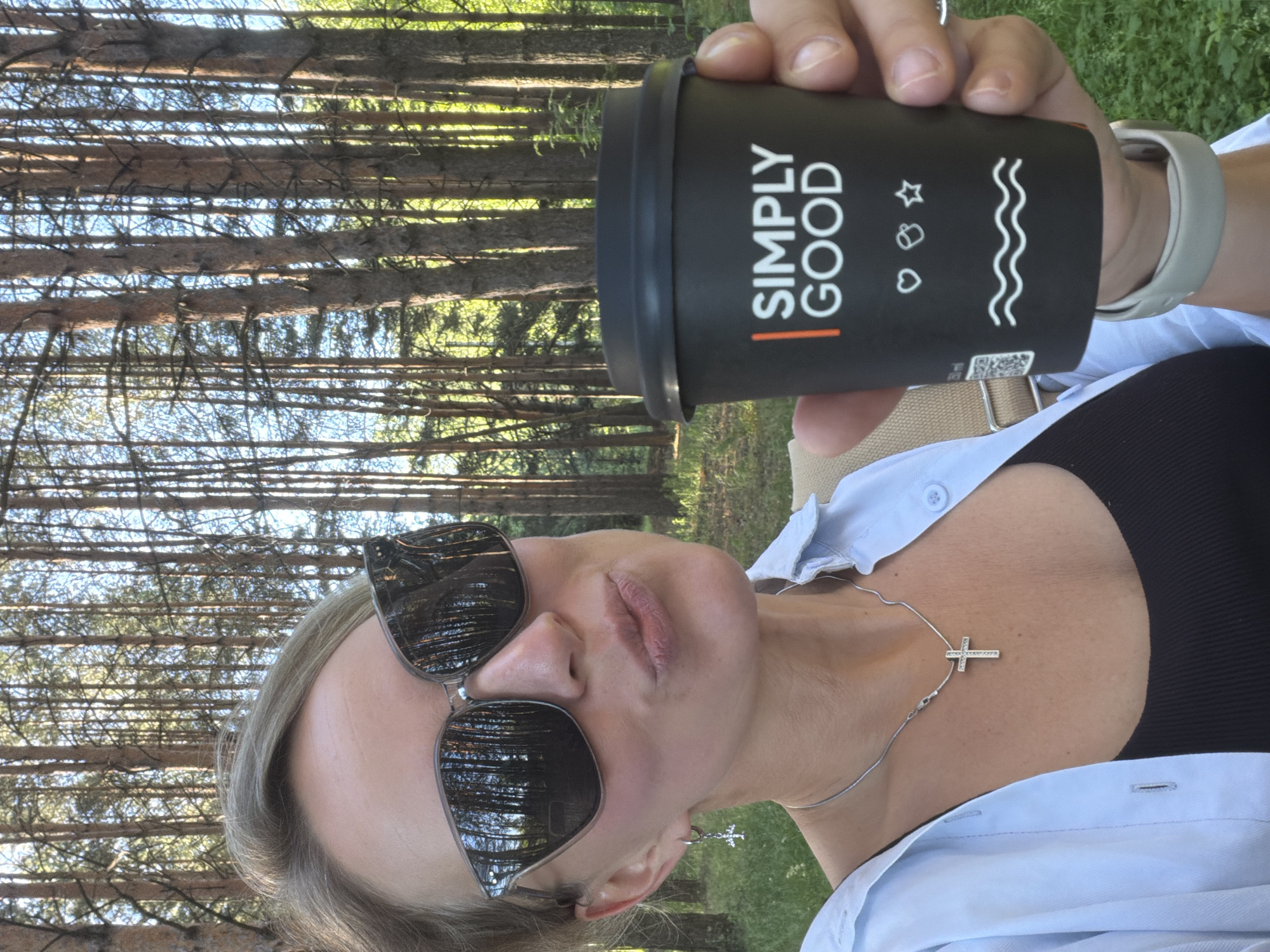 A medium shot captures a woman posing for a selfie in a sun-dappled forest, likely on the outskirts of Chaikovsky, Russia. The woman, seen from the chest up, wears large, stylish sunglasses that reflect the surrounding tall pine trees. She has blonde hair pulled back and is wearing a light blue collared shirt over a black tank top. A silver cross necklace rests on her collarbone. Her right hand holds a black disposable coffee cup with "SIMPLY GOOD" printed in white and orange text. The background is dominated by a dense stand of pine trees, with dappled sunlight filtering through the branches, suggesting it's late afternoon or early morning. The ground is covered in green grass and foliage. The overall impression is of a casual outdoor moment.