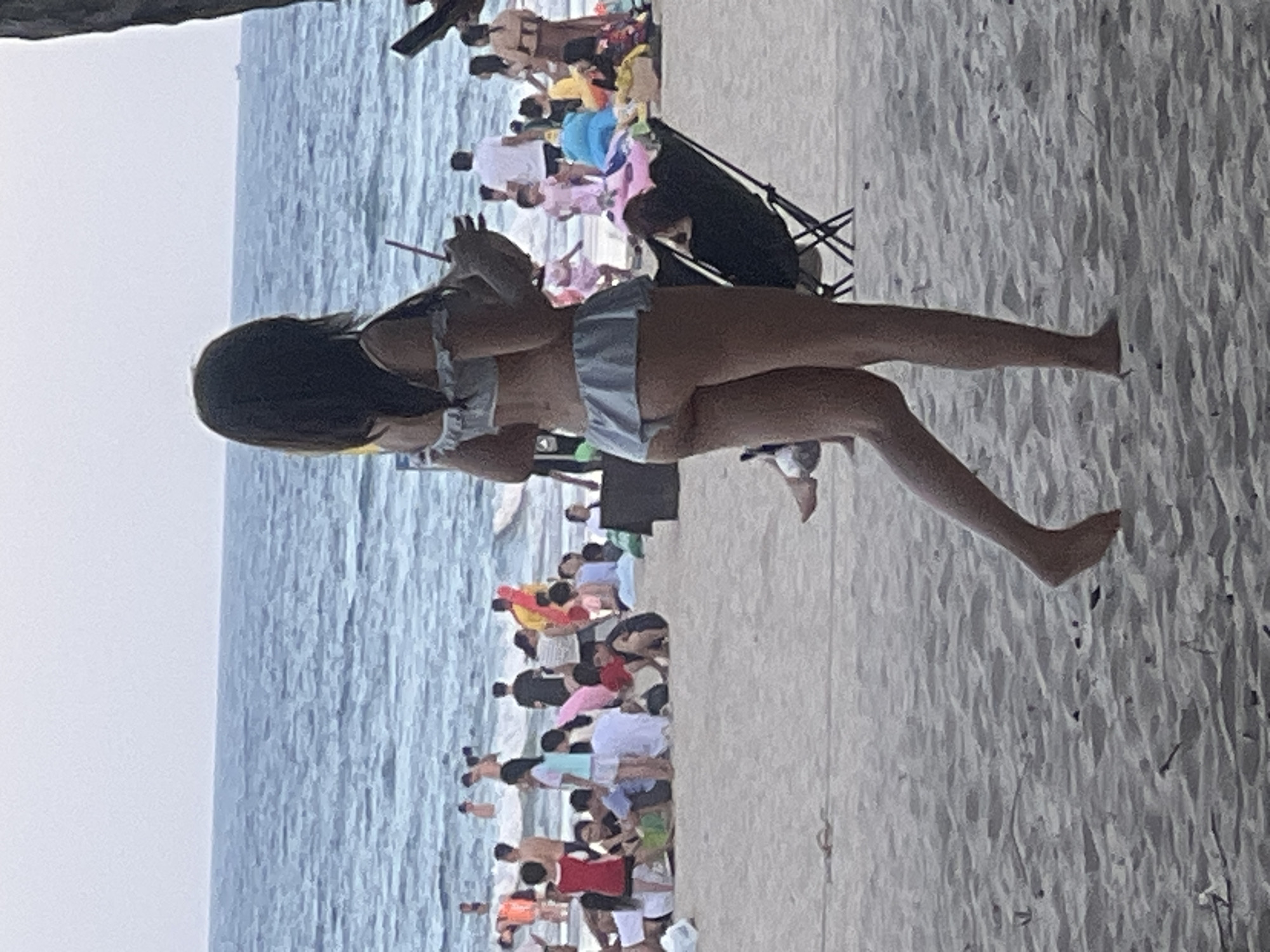 The image captures a beach scene at My Khe Beach in Đà Nẵng, Vietnam. In the foreground, a woman is seen from behind, wearing a light blue bikini with ruffled edges and holding a drink with a straw. She is walking on the sandy beach. The middle ground of the image shows a crowded beach with many people, some sitting on the sand, others standing. The ocean is visible in the background, with waves and a few people swimming or standing in the water. The sky is overcast. There are beach chairs, umbrellas, and toys scattered on the sand, indicating a leisure activity setting.