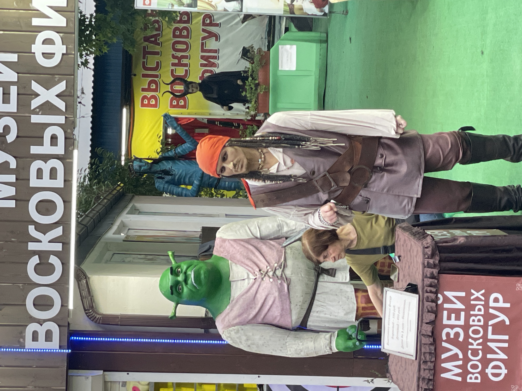 The image shows an outdoor display of wax figures in Sochi, Russia. In the foreground, a mannequin dressed as a pirate, resembling Captain Jack Sparrow, stands to the right. To the left, a large green wax figure of Shrek is positioned. Between the two figures, a woman is seated behind a table covered with a brown cloth. The table has a sign on it that reads "МУЗЕЙ ВОСКОВЫХ ФИГУР" (Museum of Wax Figures) in large white letters. Behind the wax figures, a yellow banner displays "ВЫСТАВКА ВОСКОВЫХ ФИГУР" (Wax Figure Exhibition). Other wax figures are visible in the background, including a blue figure resembling an Avatar character and a figure dressed as Maleficent with horns. The ground is covered in green artificial turf. The building behind the display has a wooden facade with large white lettering that also appears to say "МУЗЕЙ ВОСКОВЫХ ФИГУР".