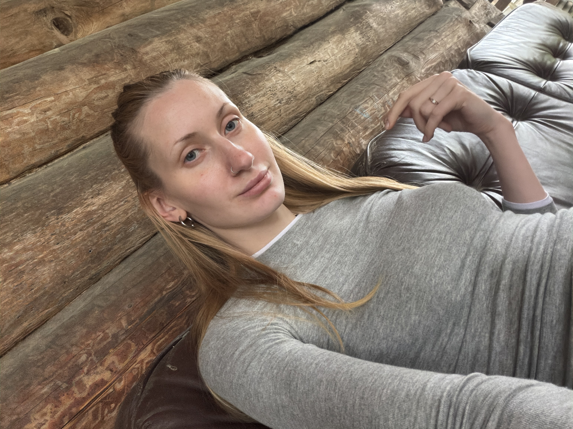 A young woman with blonde hair and blue eyes is pictured. She is wearing a grey long-sleeved shirt and a ring on her left hand. She is sitting on a tufted leather couch against a wooden wall. The location is not specified in Merkuriy, Russia.