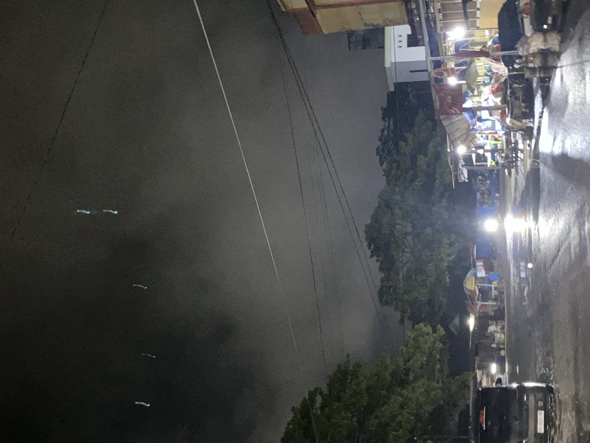 The image depicts a street scene at night in Ta', Indonesia. The sky is dark and overcast, with faint streaks of light possibly from airplanes or atmospheric phenomena. The street is wet and reflective, suggesting recent rain. On the right side of the image, there are several market stalls set up under canopies, illuminated by artificial lights. Some of these stalls appear to be selling goods, with various items and fabrics visible. In the background, there are trees and buildings. A vehicle, possibly a van or SUV, is parked on the left side of the street. The overall atmosphere is dimly lit and appears to be a bustling market area at night.