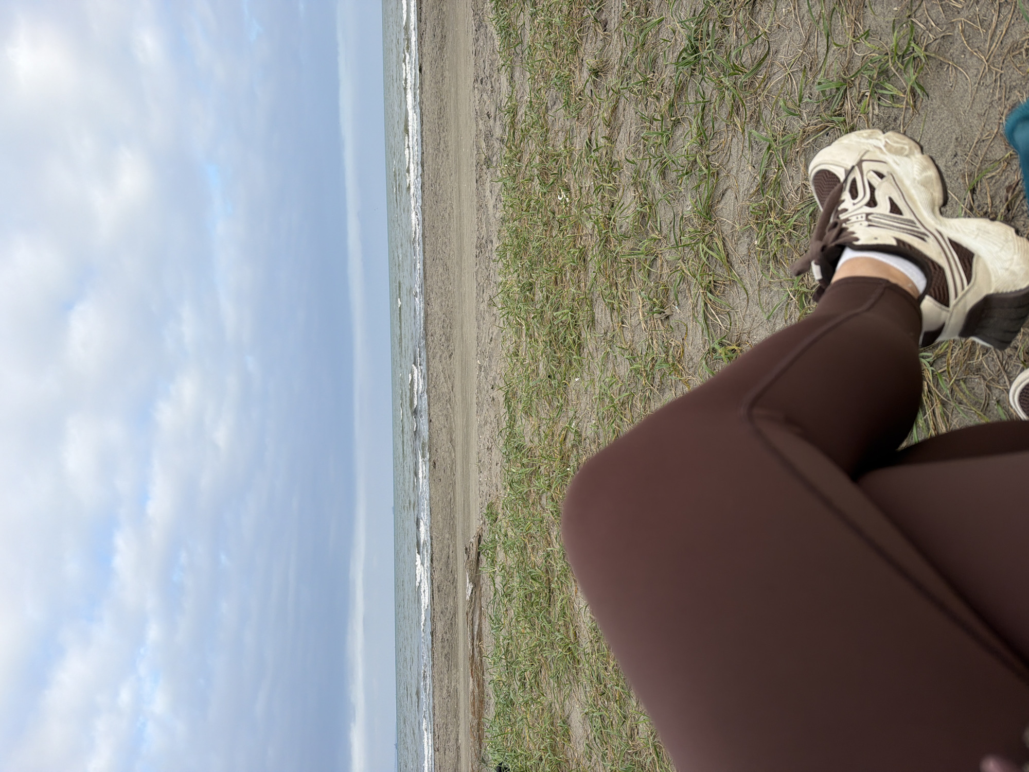 The image shows a person's legs and feet, wearing brown leggings and white and brown athletic shoes, crossed while sitting on a sandy beach. In the background, there is a calm sea with small waves under a cloudy sky. The beach has sparse patches of dry grass. The scene appears to be the coast of Sakhalin Island, potentially near Aniva, Russia, given the mention of the city.