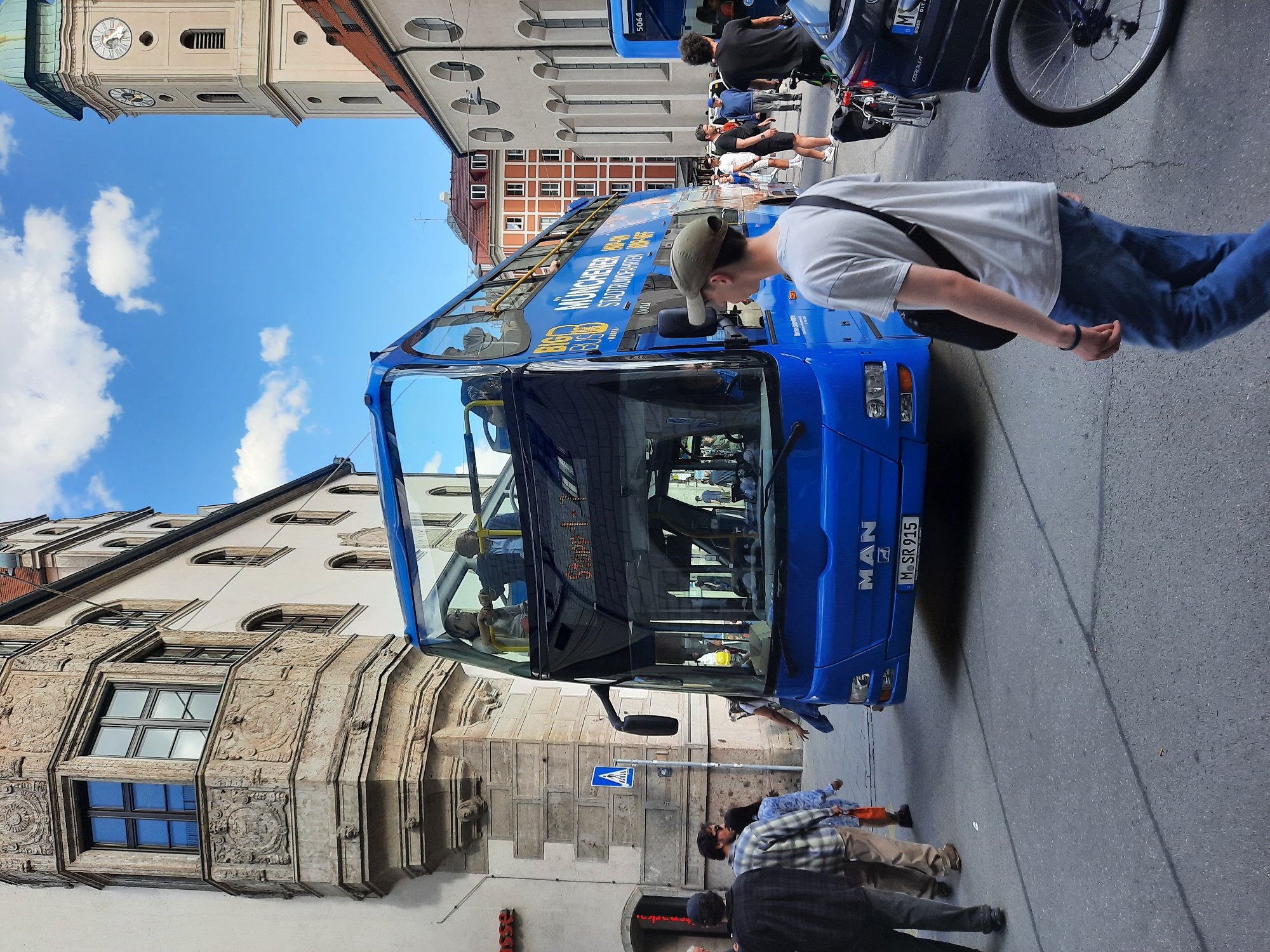 The image shows a bright, sunny day in an urban environment, likely in Europe. A prominent blue, double-decker bus, identified as a "BIG BUS MUNCHENER STADTRUNDFAHRTEN HOP-ON HOP-OFF," is the central subject, facing towards the right of the frame. The bus has "MAN" branding on the front and license plate "M SR 915." Several passengers are visible on the upper deck.
In the foreground, a young man wearing a light gray t-shirt, blue jeans, and a beige baseball cap is walking away from the camera towards the right. He has a black messenger bag slung across his body.
To the left of the bus, an ornate stone building with a bay window is visible, featuring intricate carvings. A pedestrian crossing sign is also present near this building. Further left, a glimpse of a red sign with white lettering is visible.
The background includes more buildings, one of which has a clock tower with Roman numerals. Another building to the right appears to be a church with arched windows. Other people are seen walking on the street, some with bicycles. The sky is blue with scattered white clouds. The street is paved with asphalt, and there are visible road markings and a shadow cast by the bus. A dark-colored car is parked on the right side of the street, with a bicycle wheel visible in the foreground.