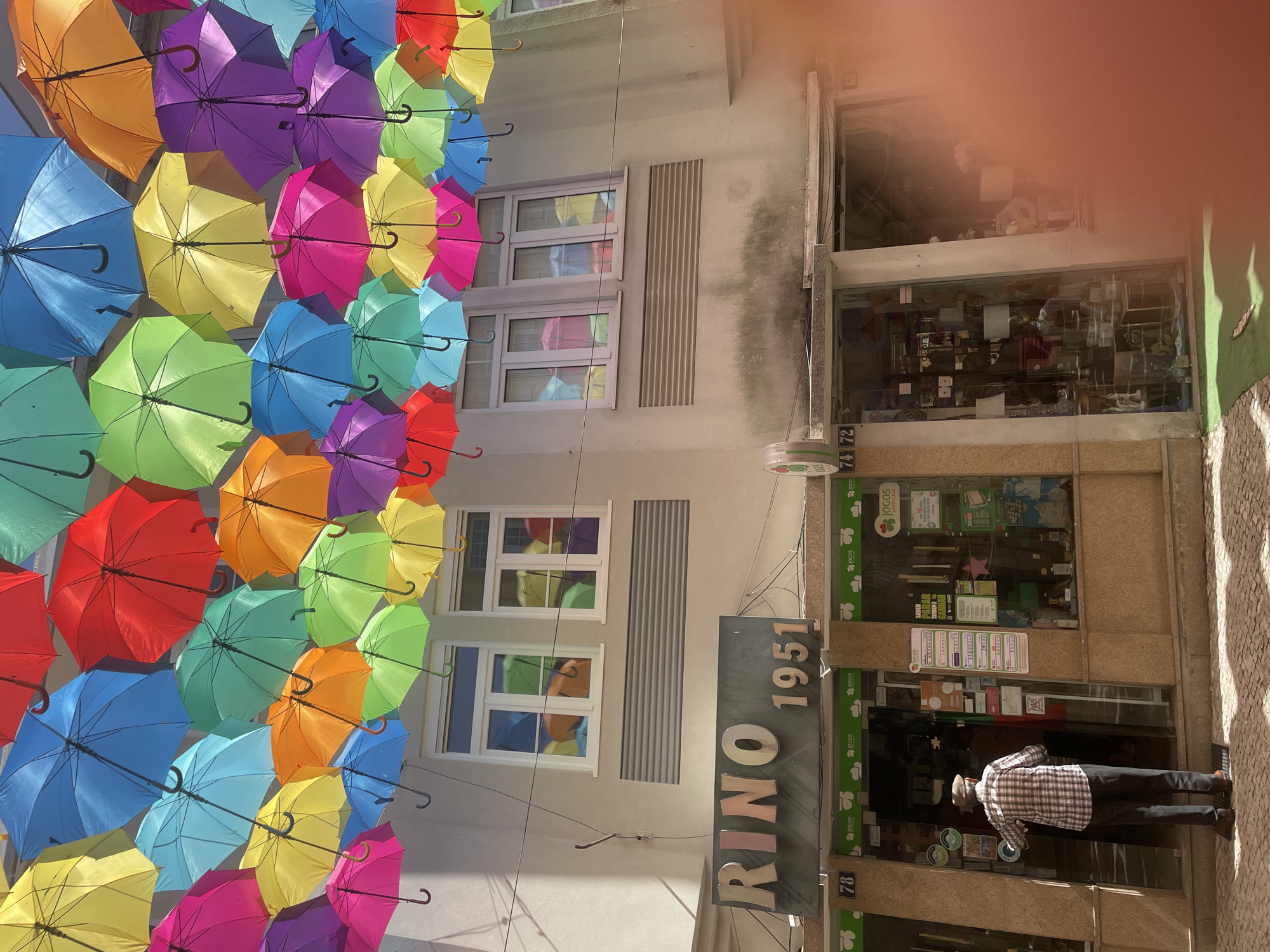 This image captures a vibrant street scene in Águeda, Portugal, likely during daylight hours given the bright sunlight casting shadows. The most striking feature is an overhead installation of colorful, open umbrellas suspended across a narrow street, creating a lively and festive canopy. The umbrellas are arranged in a dense, cascading pattern, showcasing a rainbow of hues including yellow, blue, green, orange, pink, purple, and red.
Below the umbrellas, a two-story building is visible with its facade partially obscured. The building has several windows, and the reflection of the colorful umbrellas can be seen in the glass. A prominent sign with metallic letters reads "RINO 1951," suggesting the name and establishment year of a business. Further down, the storefront of a shop is visible, with a glass facade displaying various items and signage including "JOGOS SANTACASA" and numerical markers like "78," "74," and "72."
A person, seen from behind, is standing in front of the shop entrance, appearing to interact with something inside or looking at the displays. They are wearing a plaid shirt and dark pants. The pavement is made of cobblestones, adding to the traditional streetscape feel. The overall atmosphere is cheerful and visually engaging, highlighted by the abundance of color and the unique umbrella installation.