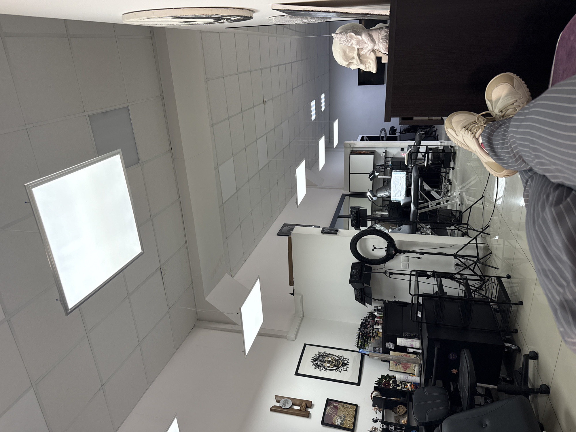 The image shows the interior of a tattoo parlor, potentially located in Rostov-on-Don, Russia, based on contextual clues if available but not verifiable from the image alone. The room features a grid-patterned ceiling with recessed lighting panels, some of which are illuminated. A white skull sculpture is visible on a dark surface in the upper part of the image. Tattoo equipment, including a ring light, work stations with multiple screens, and shelves stocked with tattoo inks and supplies, are arranged throughout the room. Black rolling carts and a tattoo artist's chair are present. Artwork, including framed pieces and a mandala-style skull design, is displayed on the walls. A person is partially visible in the foreground, seated and wearing tan-colored sneakers and a striped shirt. The floor appears to be tiled. The environment suggests a professional and sterile setting for tattoo artistry.