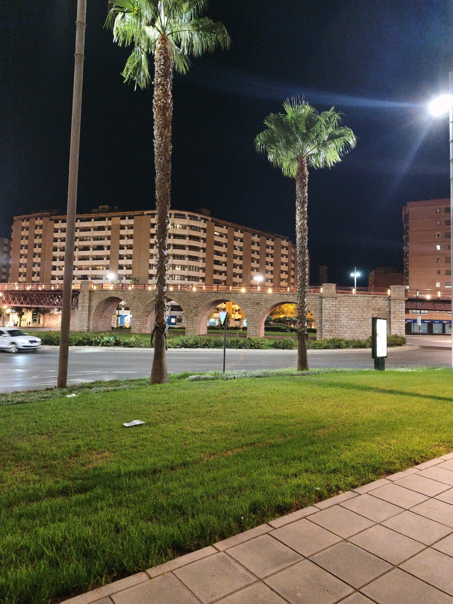The image depicts an urban night scene in Almeria, Spain, featuring a large, multi-story building with many balconies and windows. In the foreground, a well-maintained patch of green grass lines a paved walkway. Two tall palm trees stand prominently on either side of the frame, with their fronds illuminated by streetlights. A stone bridge with multiple arches crosses the scene in the mid-ground, its structure adorned with lights. A road runs in front of the bridge, and a white car is captured in motion, its headlights on. The sky is dark, indicating nighttime. There are no visible people in the image, and no discernible actions are taking place beyond the movement of the car. The overall environment is one of a well-lit, quiet urban area at night.